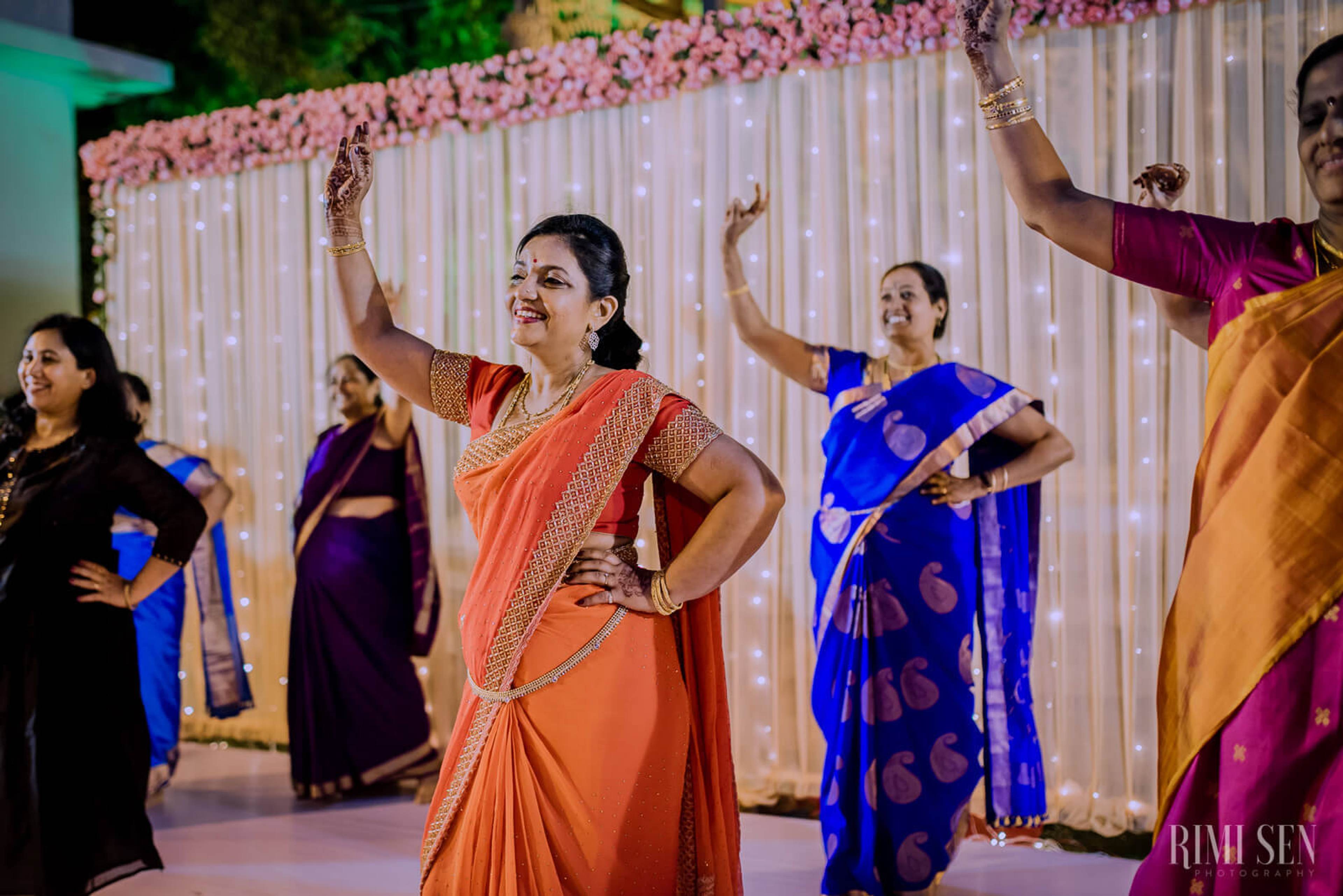 Woman in orange saree dancing with arms raised at Bangalore wedding celebration, pink floral backdrop
