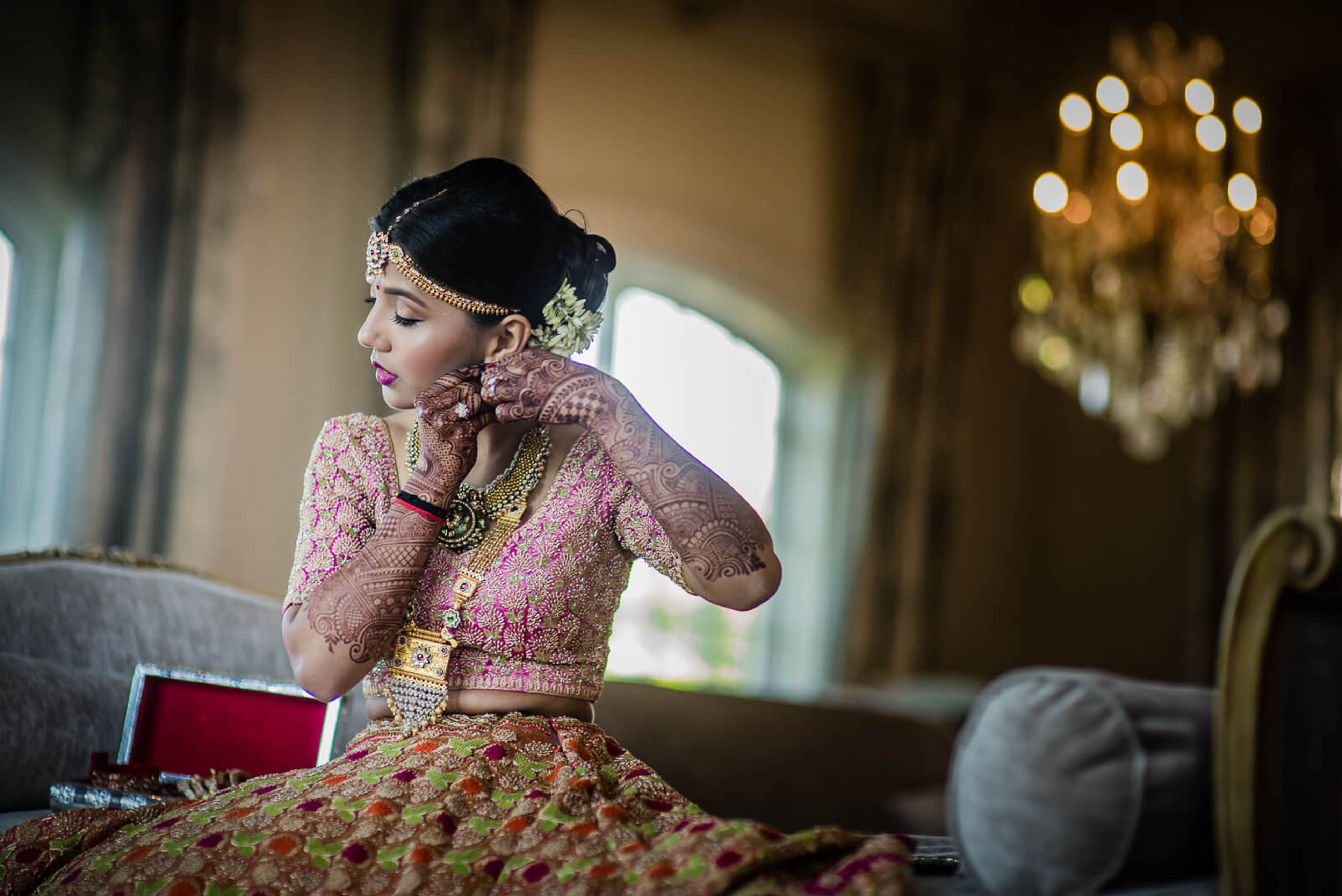 Barkha in ornate pink and gold traditional attire, jewelry, and headpiece, posing thoughtfully indoors under chandelier