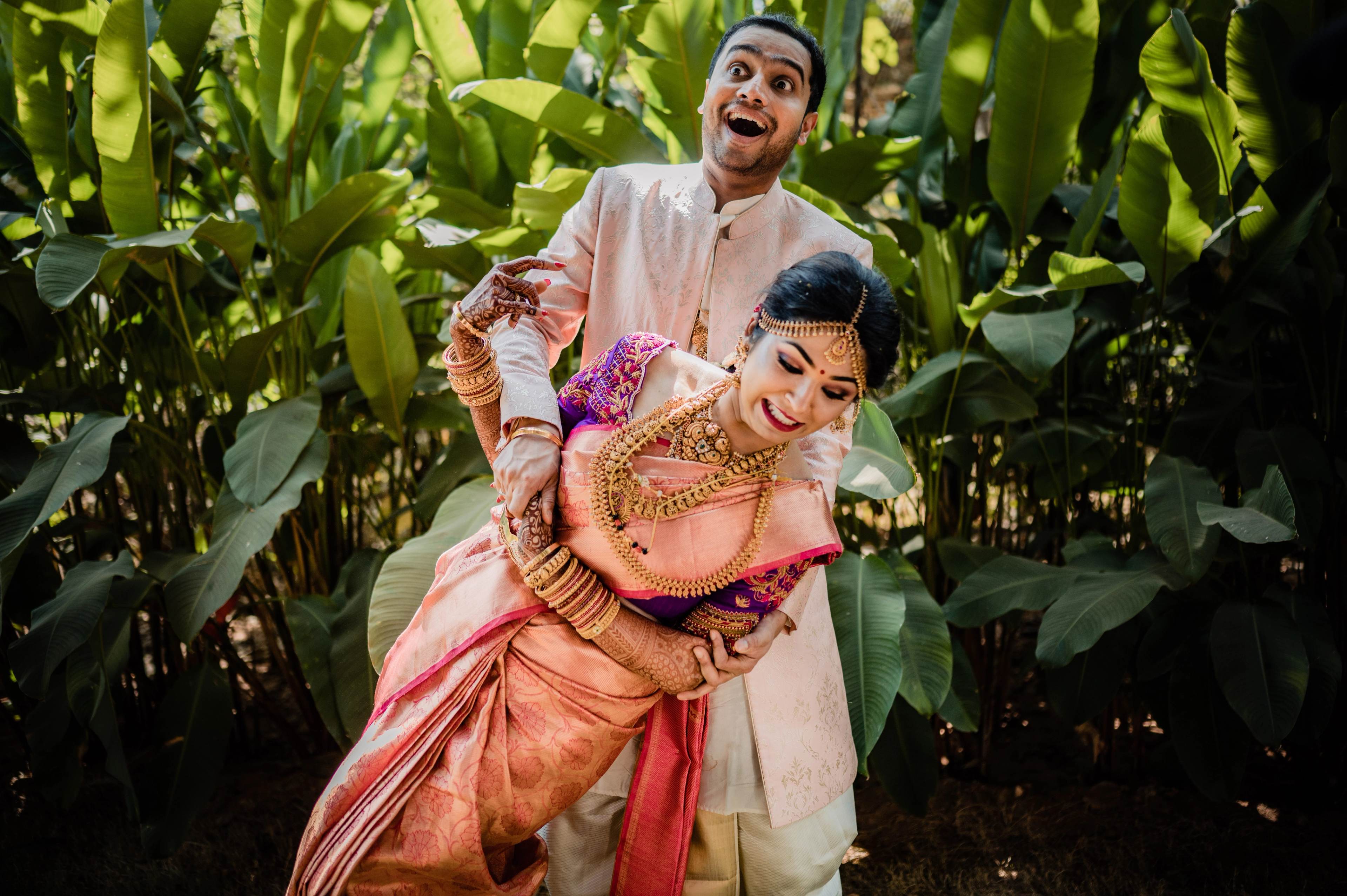 Groom in cream sherwani playfully lifting bride in coral silk saree amid lush green foliage
