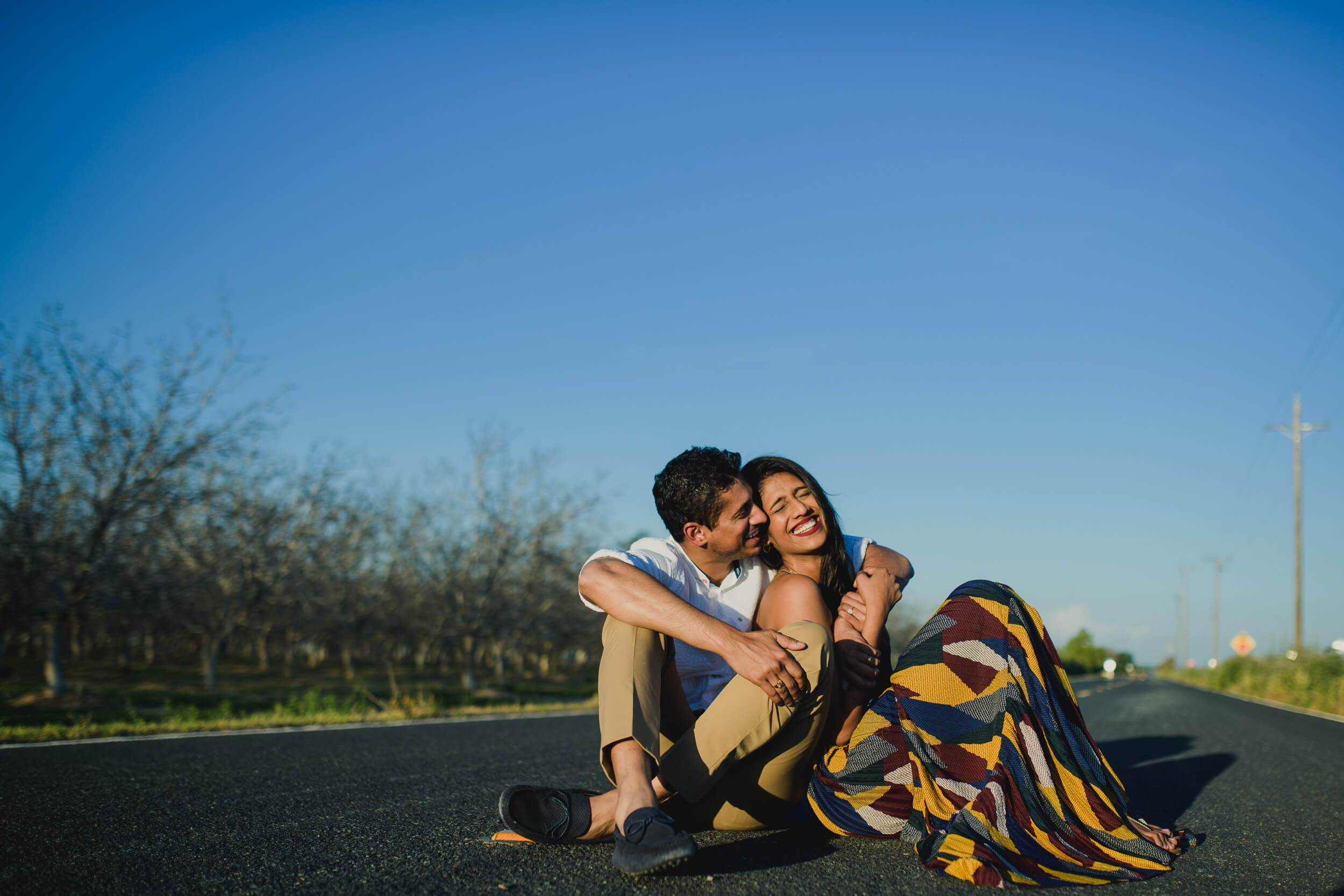 Anu and Raghu sit embracing on a rural California road under clear blue sky, orchards behind them