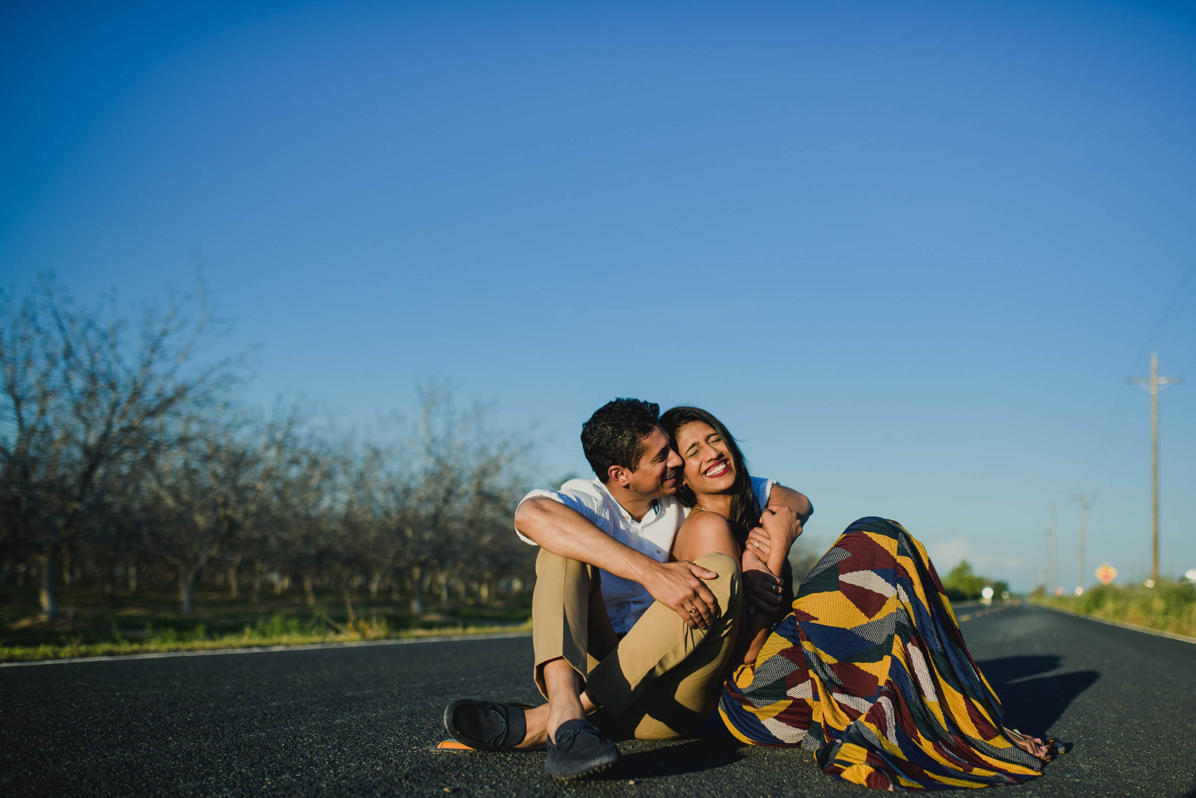 Anu and Raghu sit embracing on a rural California road under clear blue sky, orchards behind them