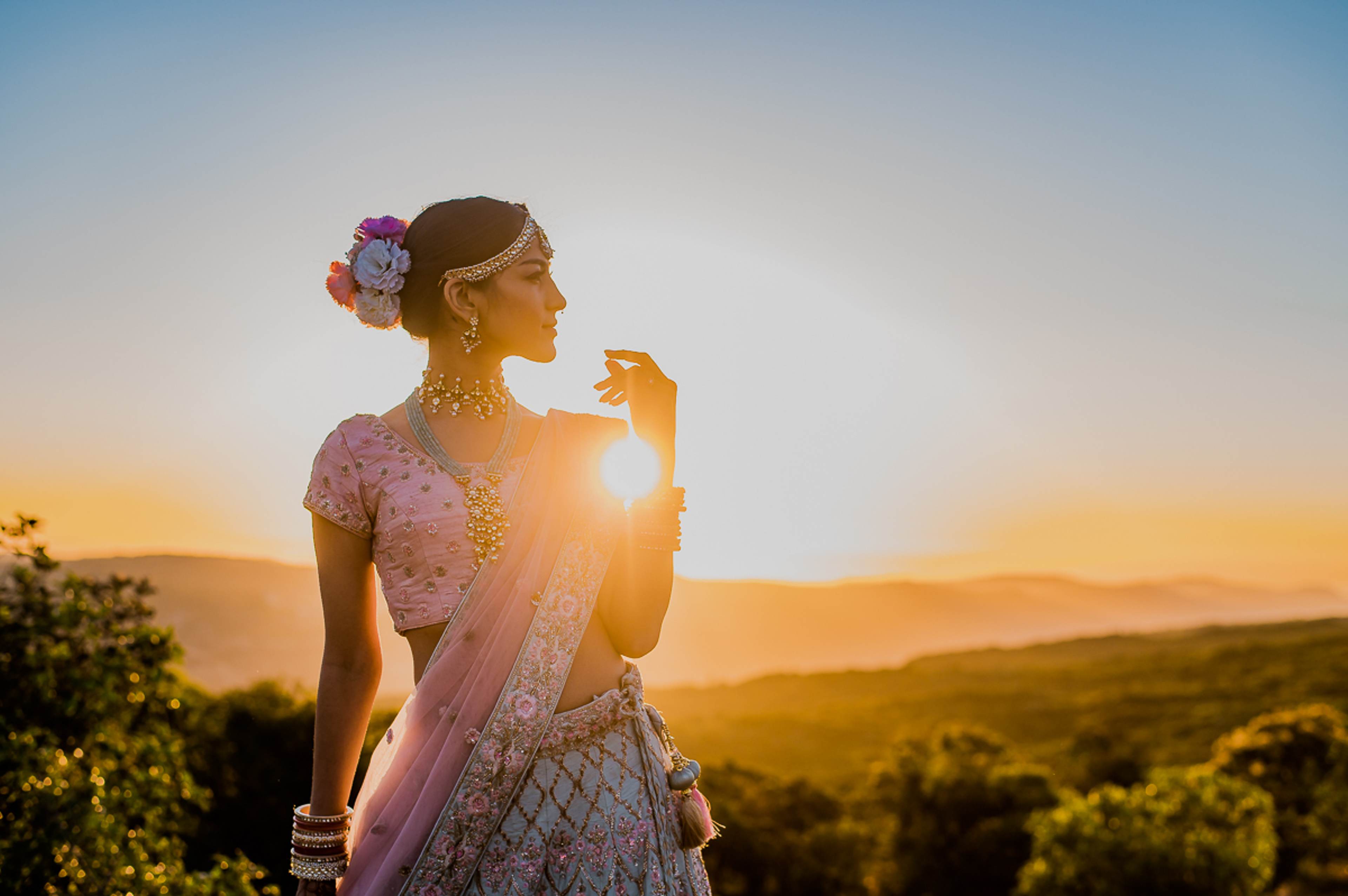 Woman in pink traditional attire with floral hair piece, backlit by golden sunset over rolling hills