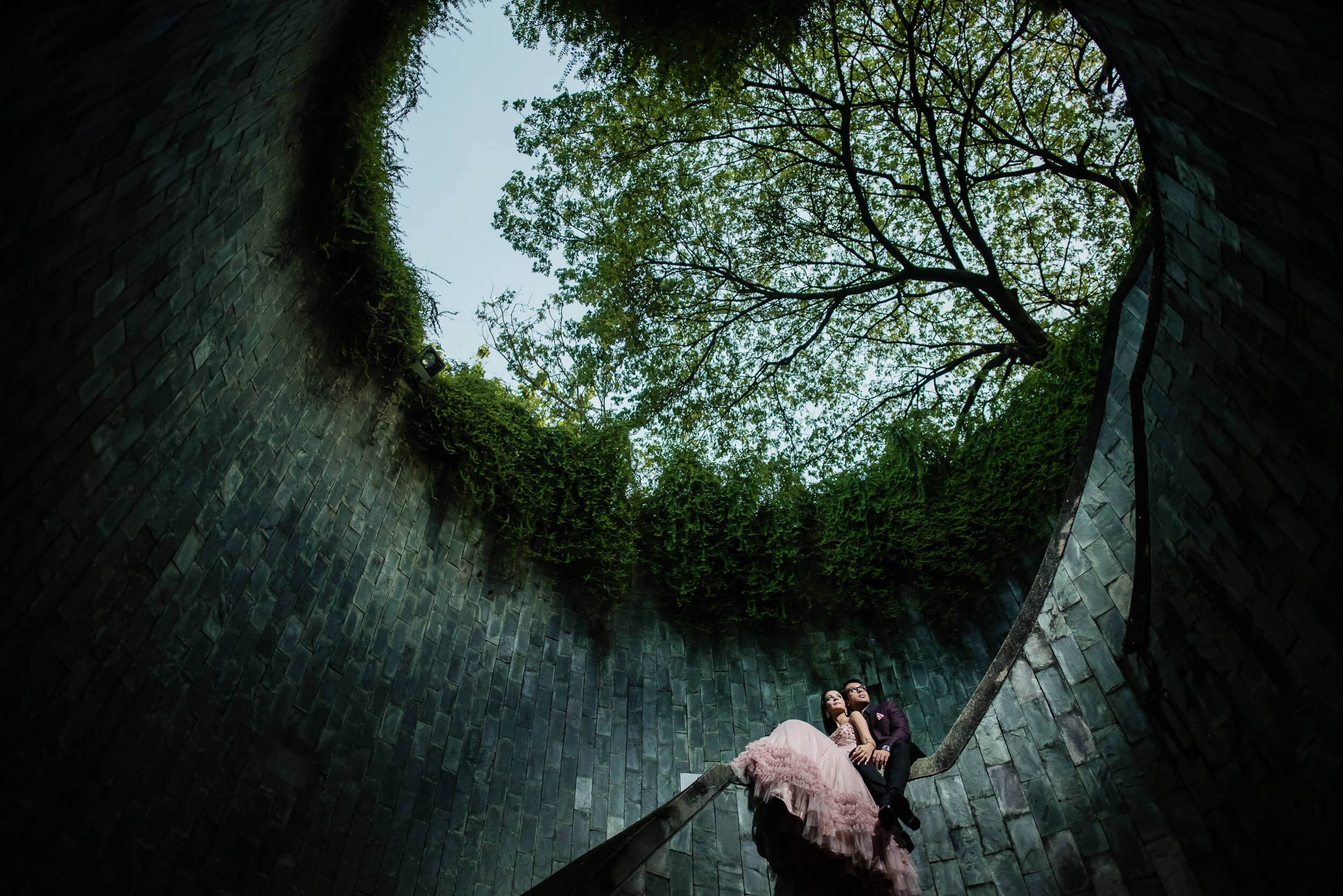 Yash and Disha stand in a curved brick archway with ivy and trees overhead in Singapore by Rimi Sen