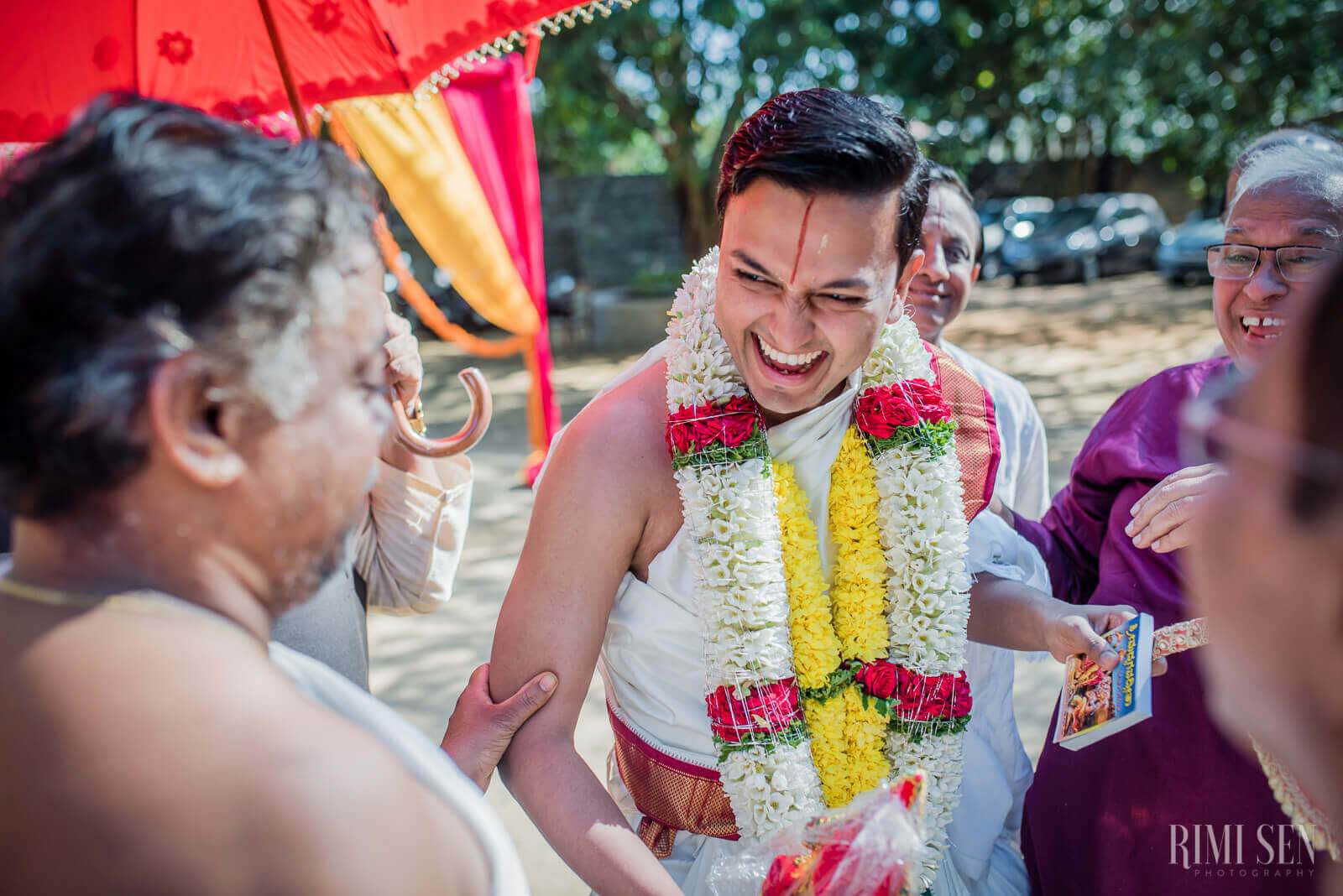 Aniket smiles brightly wearing a white shirt and red and yellow flower garland, surrounded by family at an outdoor celebration in Bangalore