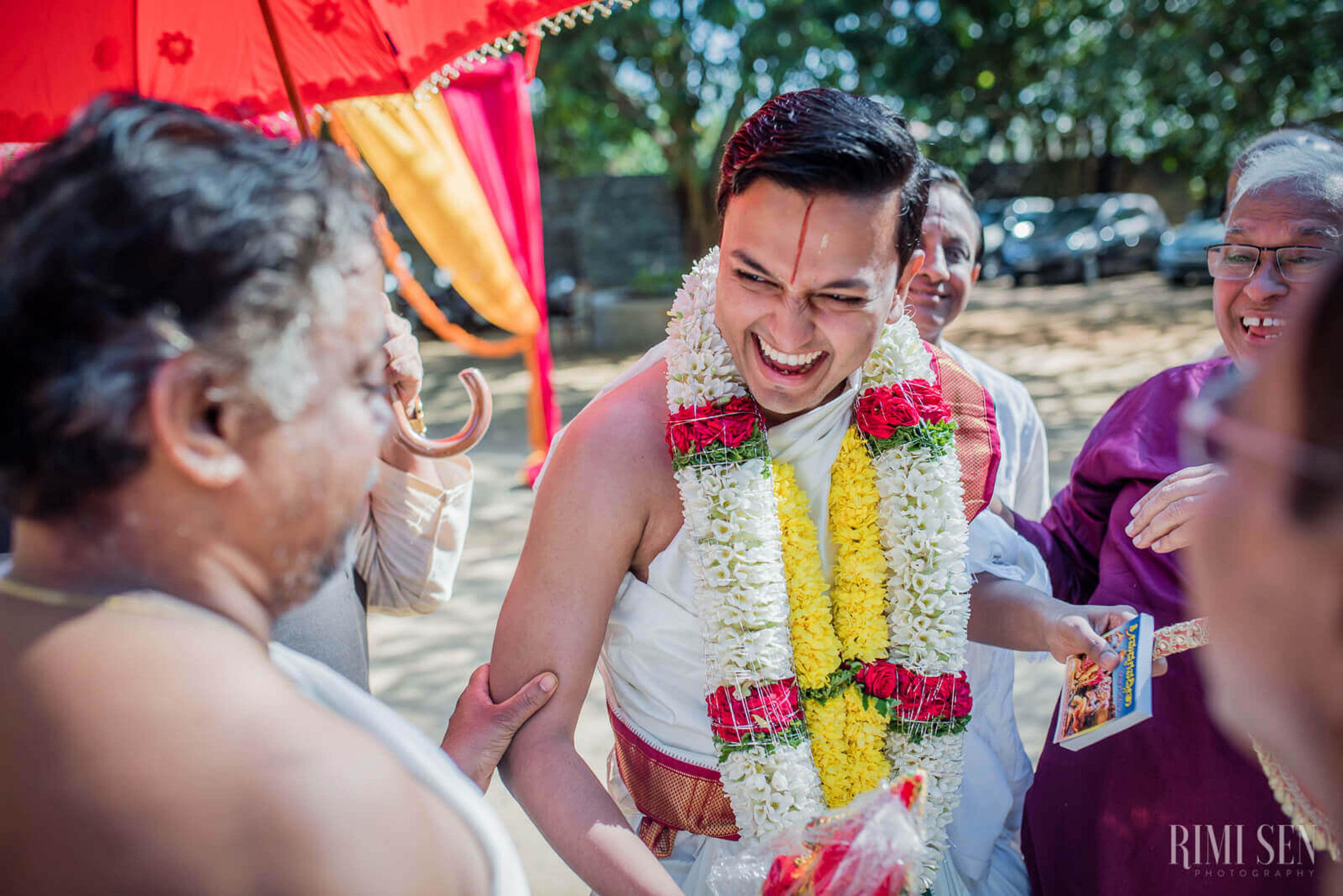 Aniket smiles brightly wearing a white shirt and red and yellow flower garland, surrounded by family at an outdoor celebration in Bangalore