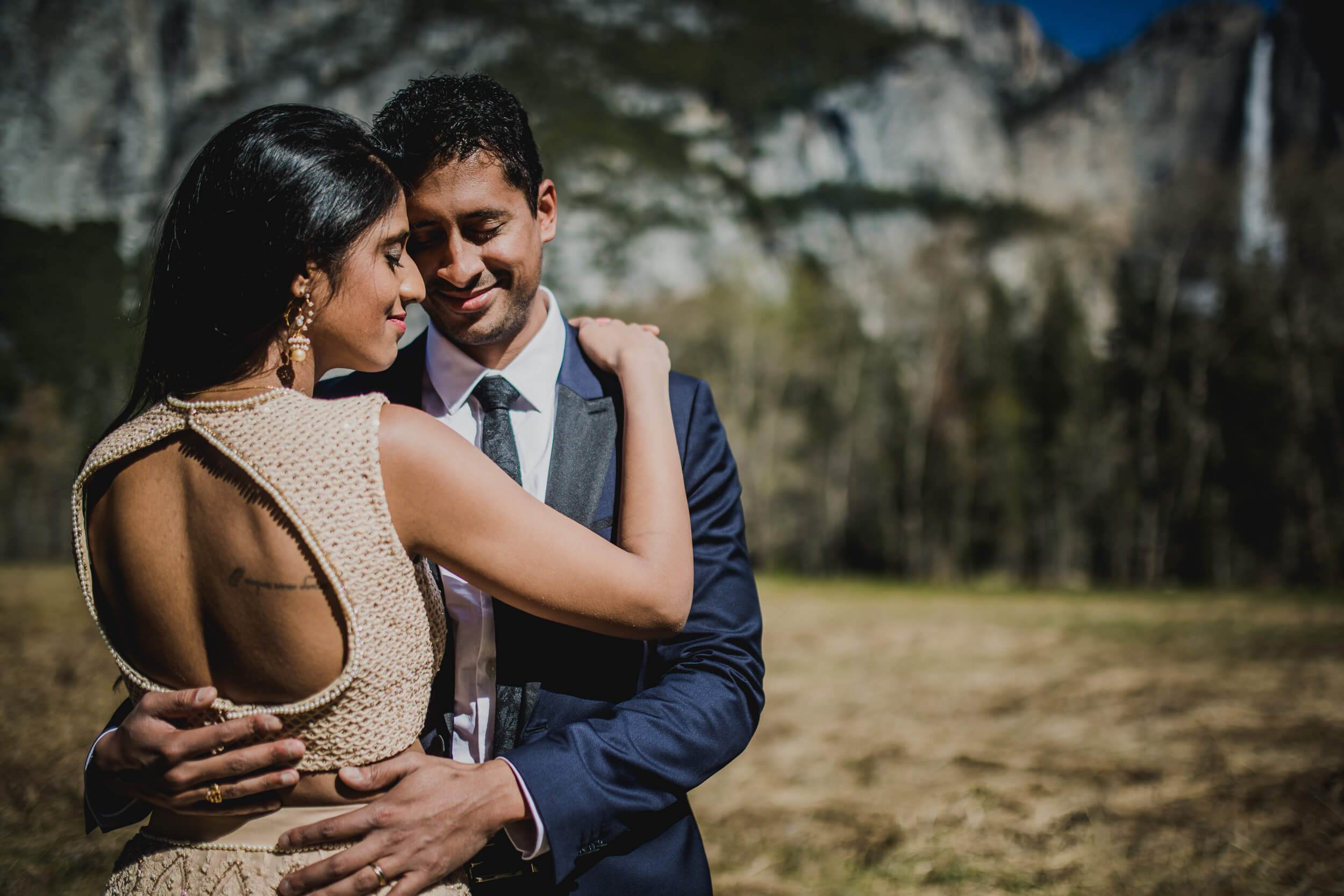 Anu and Raghu embrace in a sun-drenched California meadow with granite cliffs and forest behind