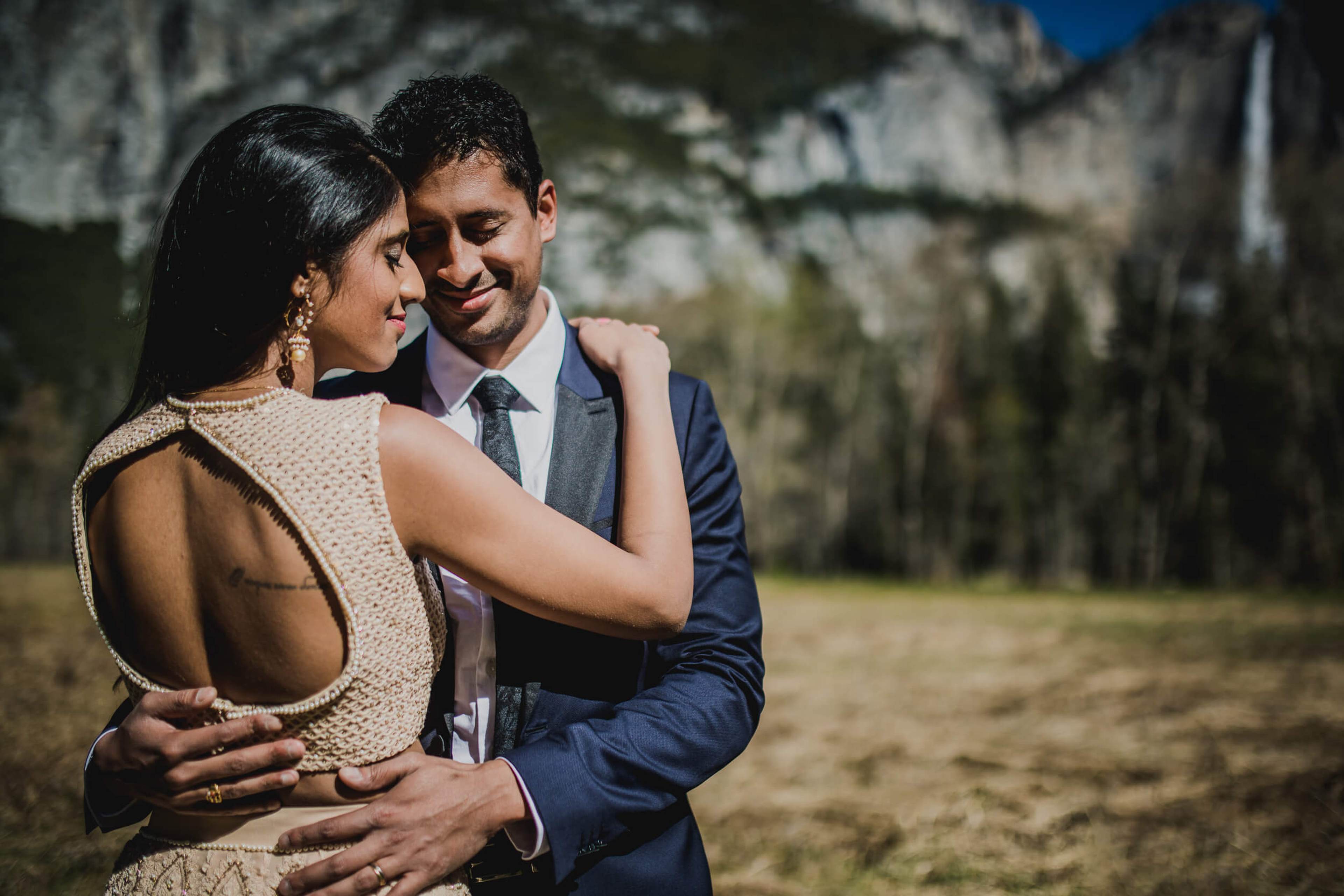 Anu and Raghu embrace in a sun-drenched California meadow with granite cliffs and forest behind