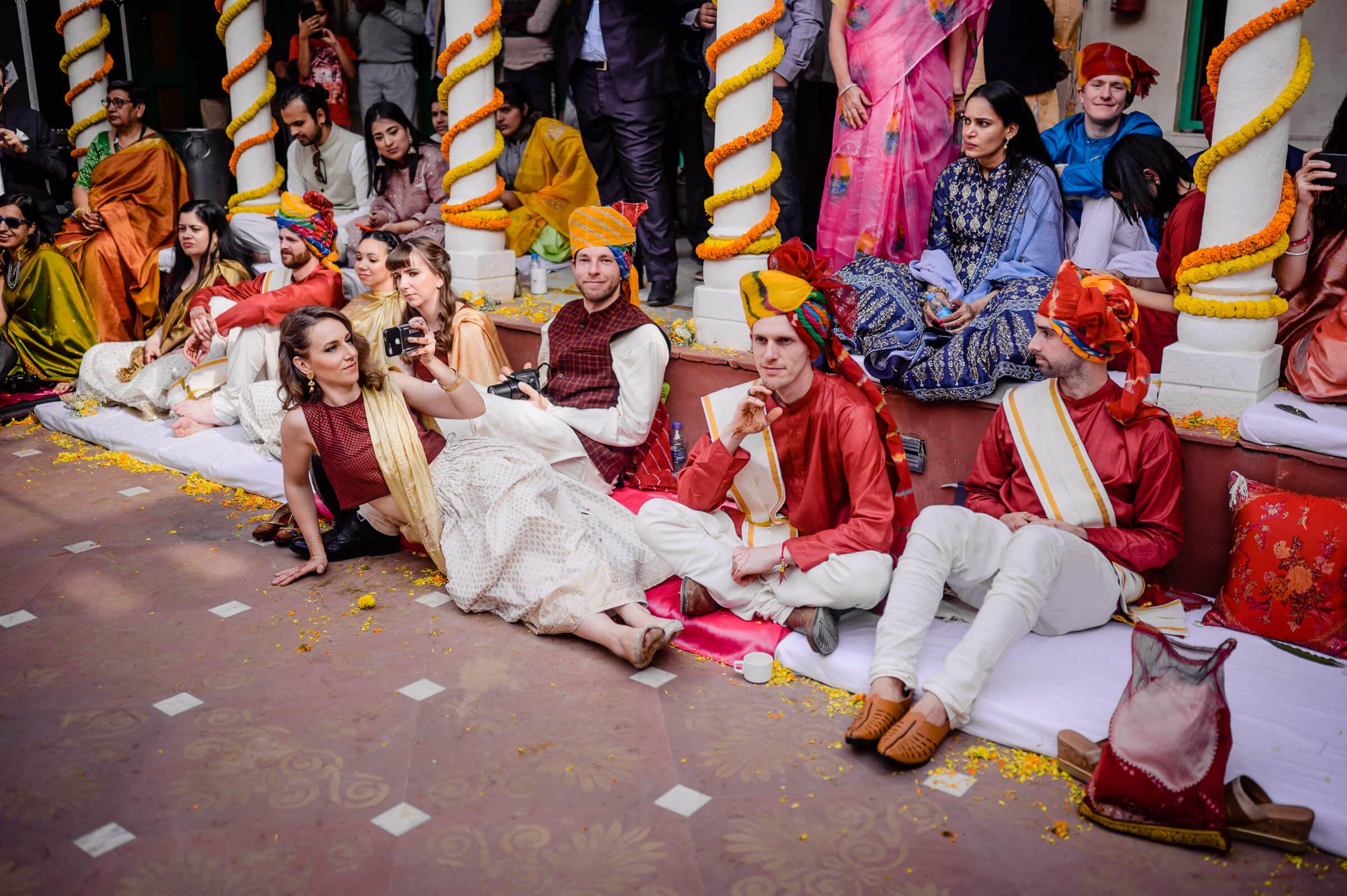 Maitrayee and Eric sit on ornate floor surrounded by colorful marigold garlands and guests in traditional Indian wedding attire