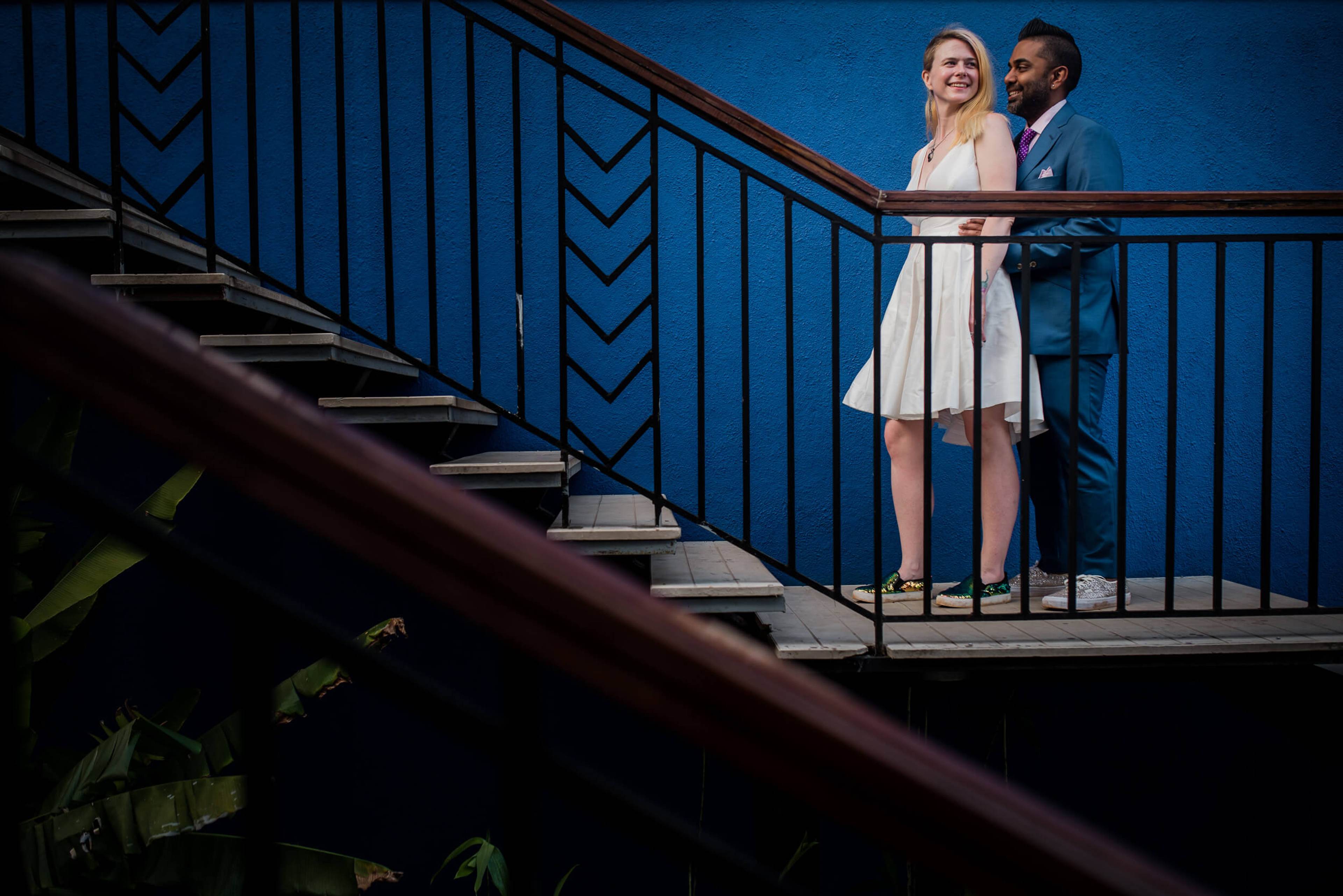 Sarah and Ravin stand together on a blue-railed balcony in Goa, smiling in natural light