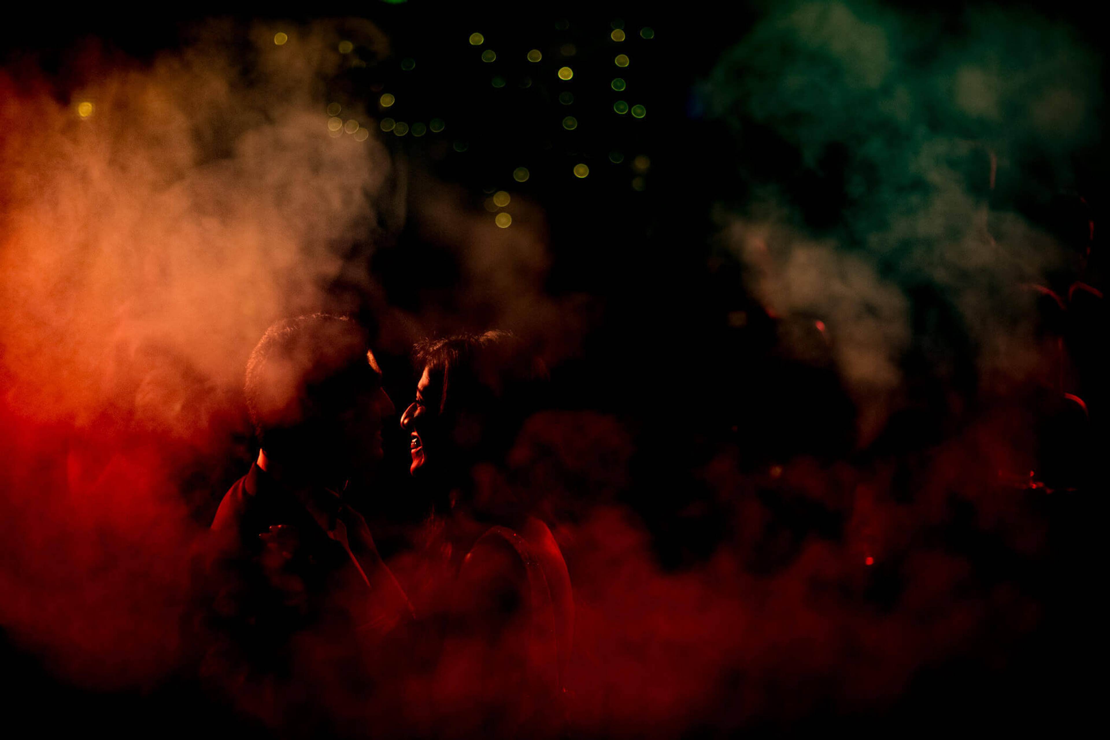 Anu and Raghu silhouetted in vibrant red and green powder clouds at night