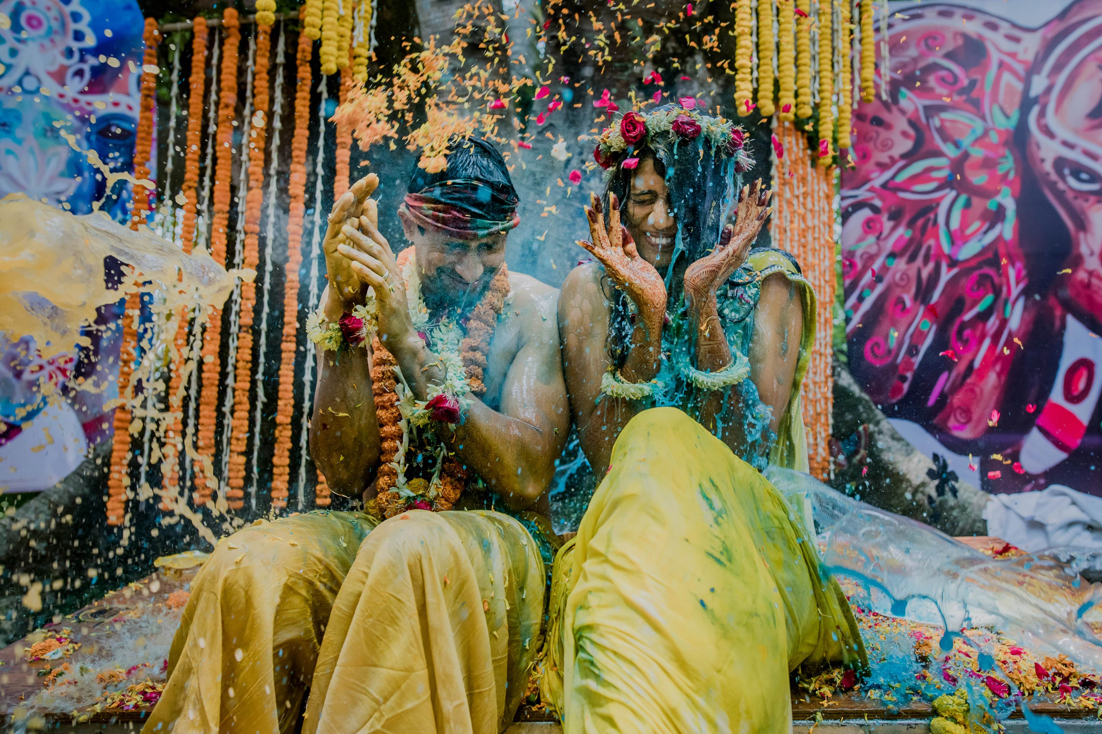 Couple covered in colorful powder and flower petals during Holi celebration with vibrant decorated backdrop