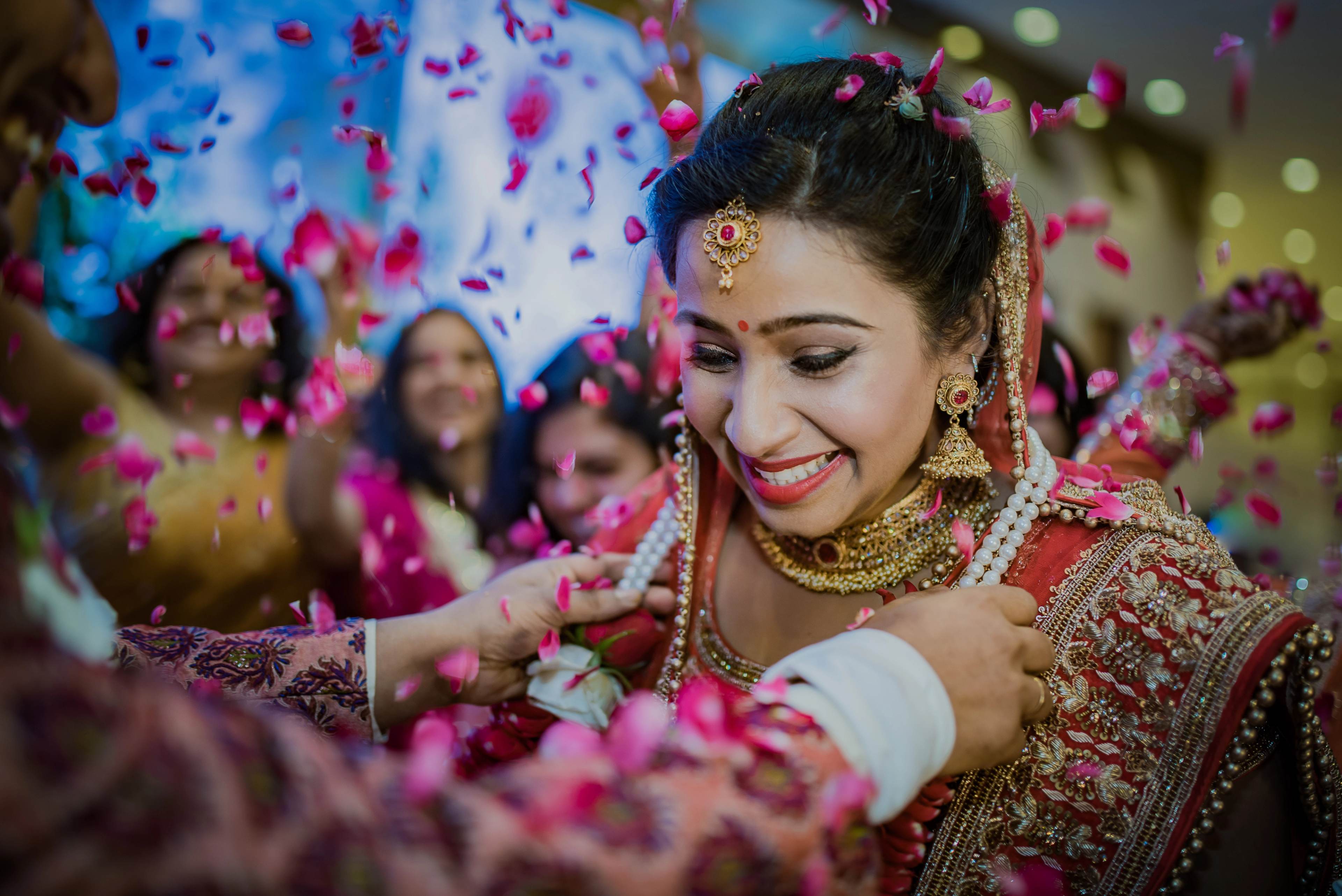 Woman in ornate red and gold wedding attire smiling as pink flower petals fall, celebrating with guests