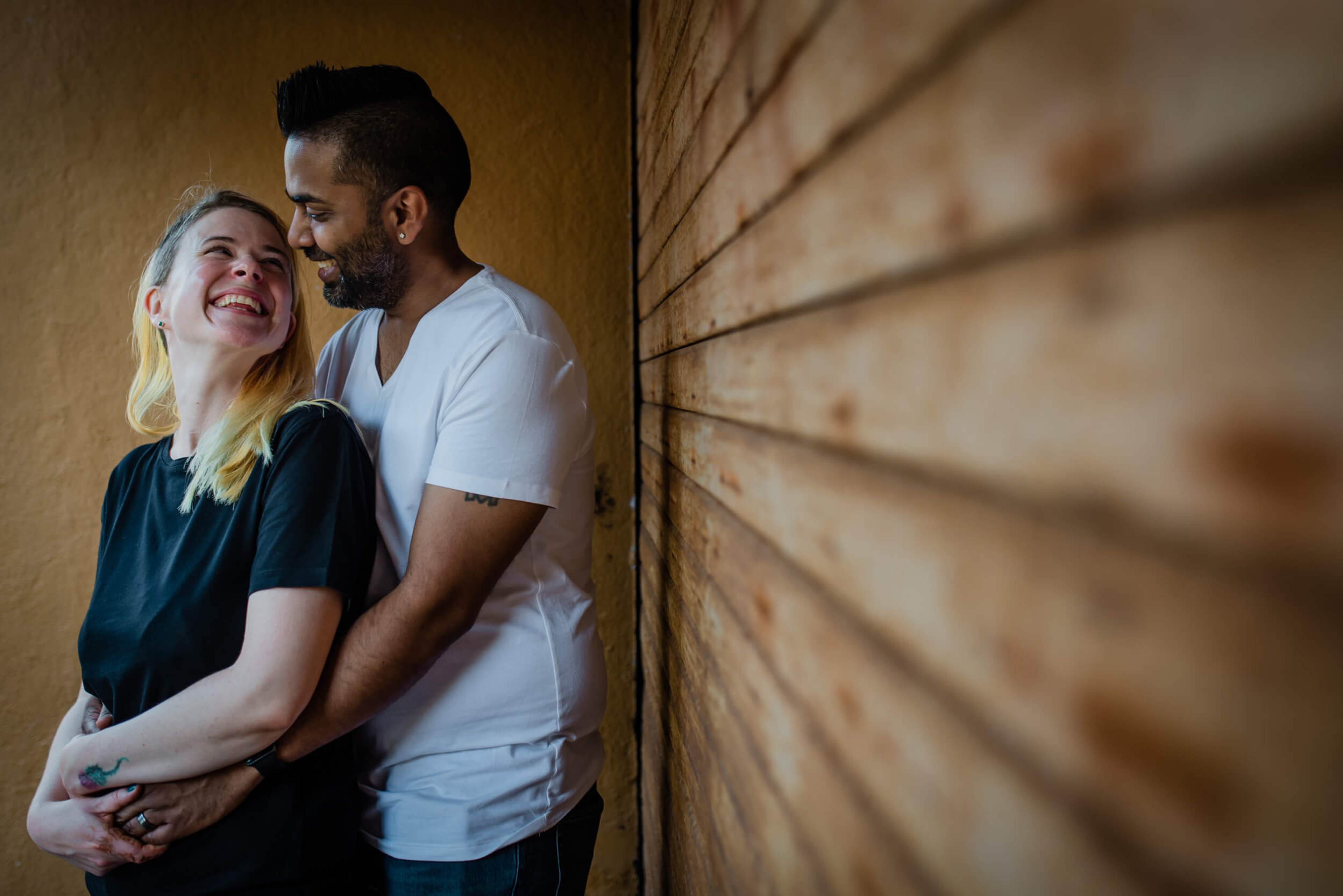 Sarah and Ravin embrace against a textured brick wall, smiling in warm golden light