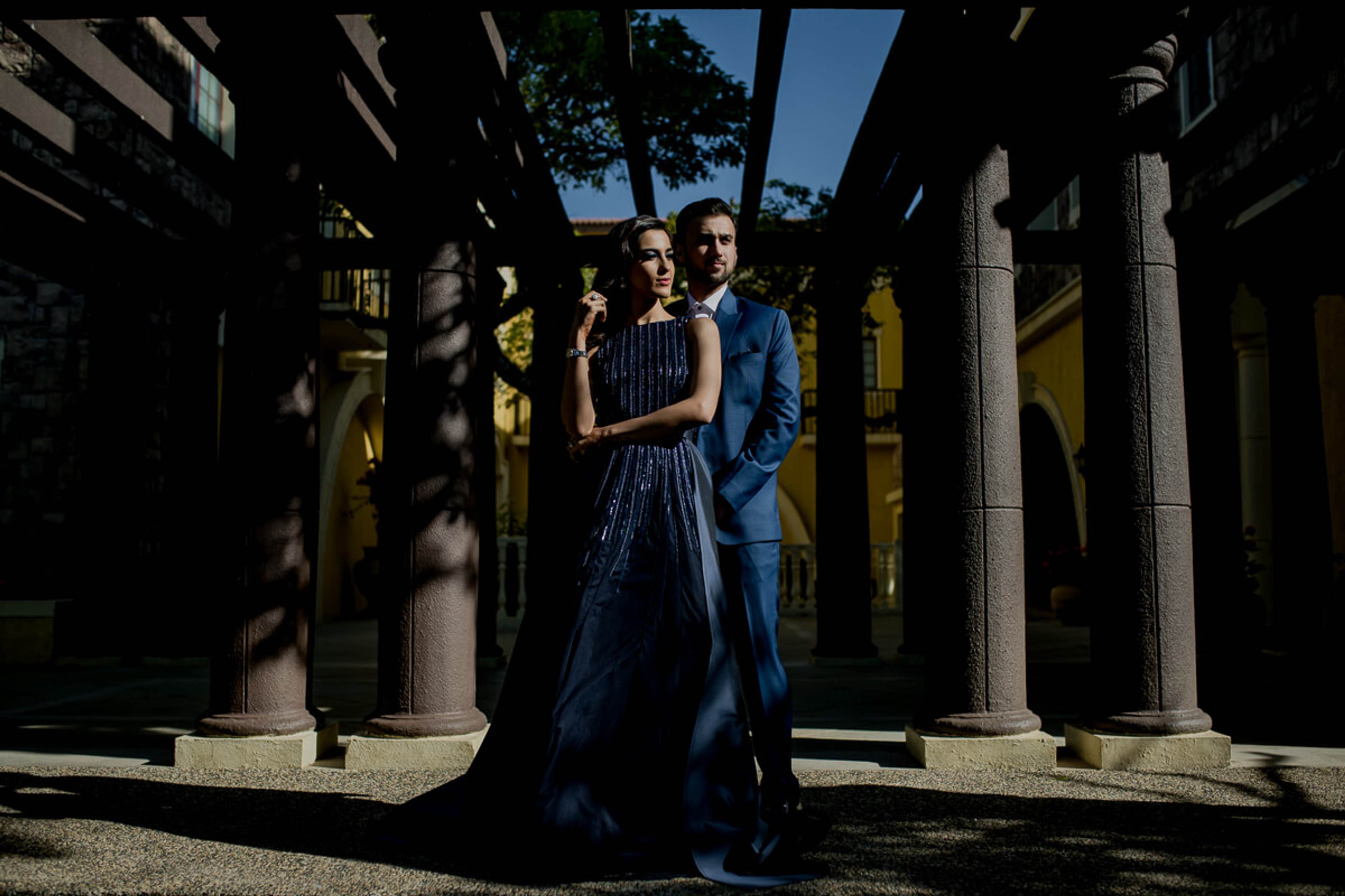 Shreya and Yash stand between stone columns in dappled sunlight, dressed in coordinated blue formal wear