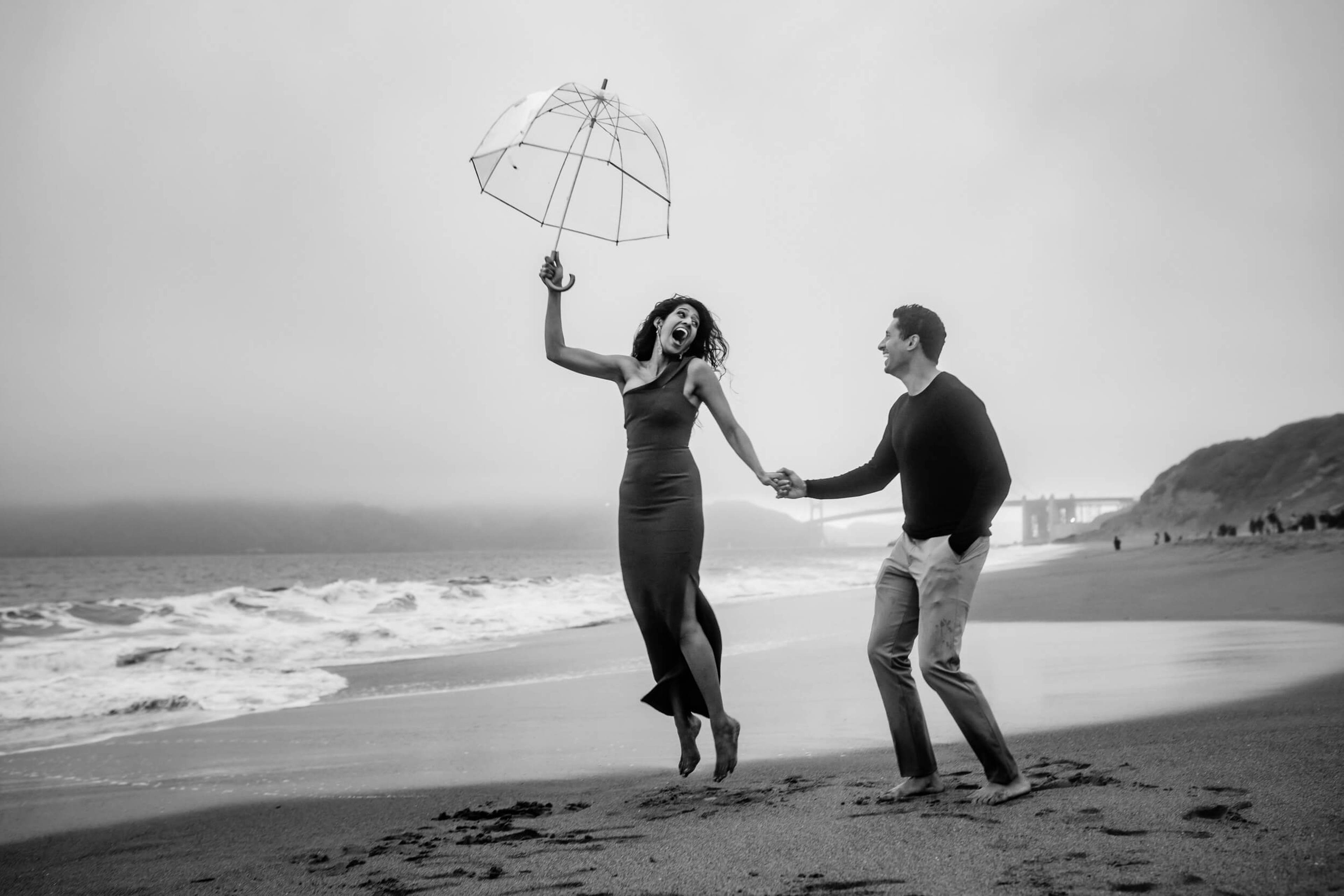 Anu and Raghu hold hands on a California beach, she jumping joyfully with a clear umbrella in black and white