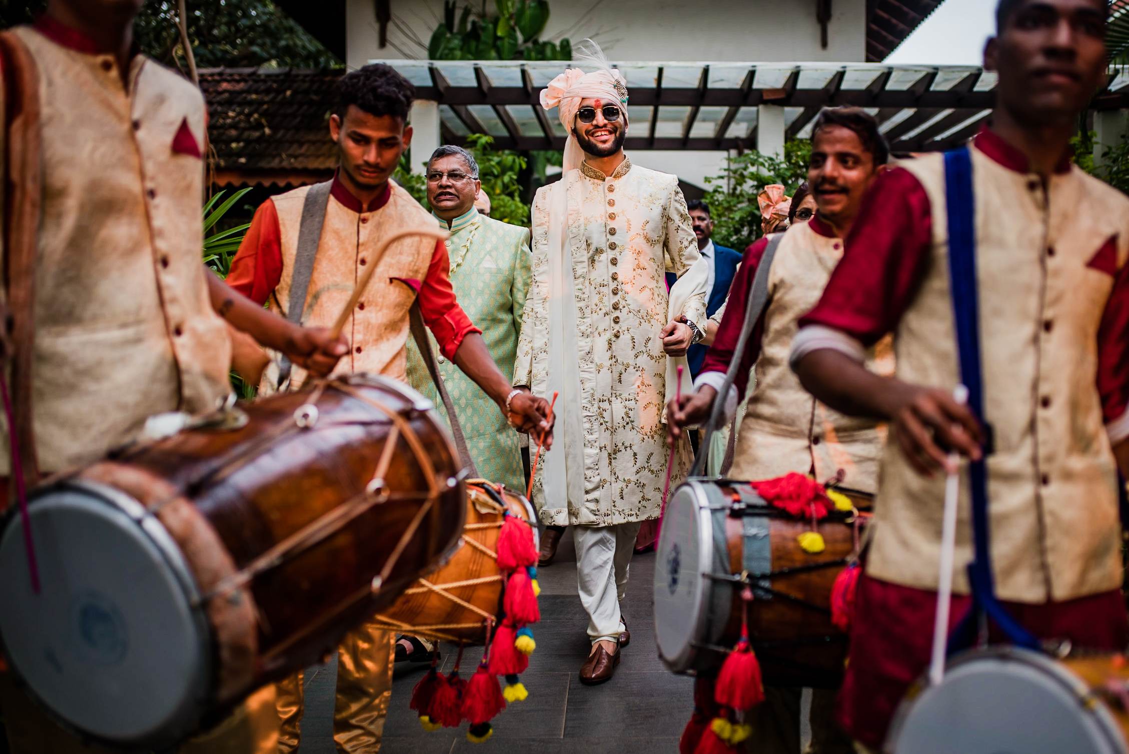 Avani and Srikant walk through a festive garden ceremony in Goa, surrounded by drummers in traditional attire