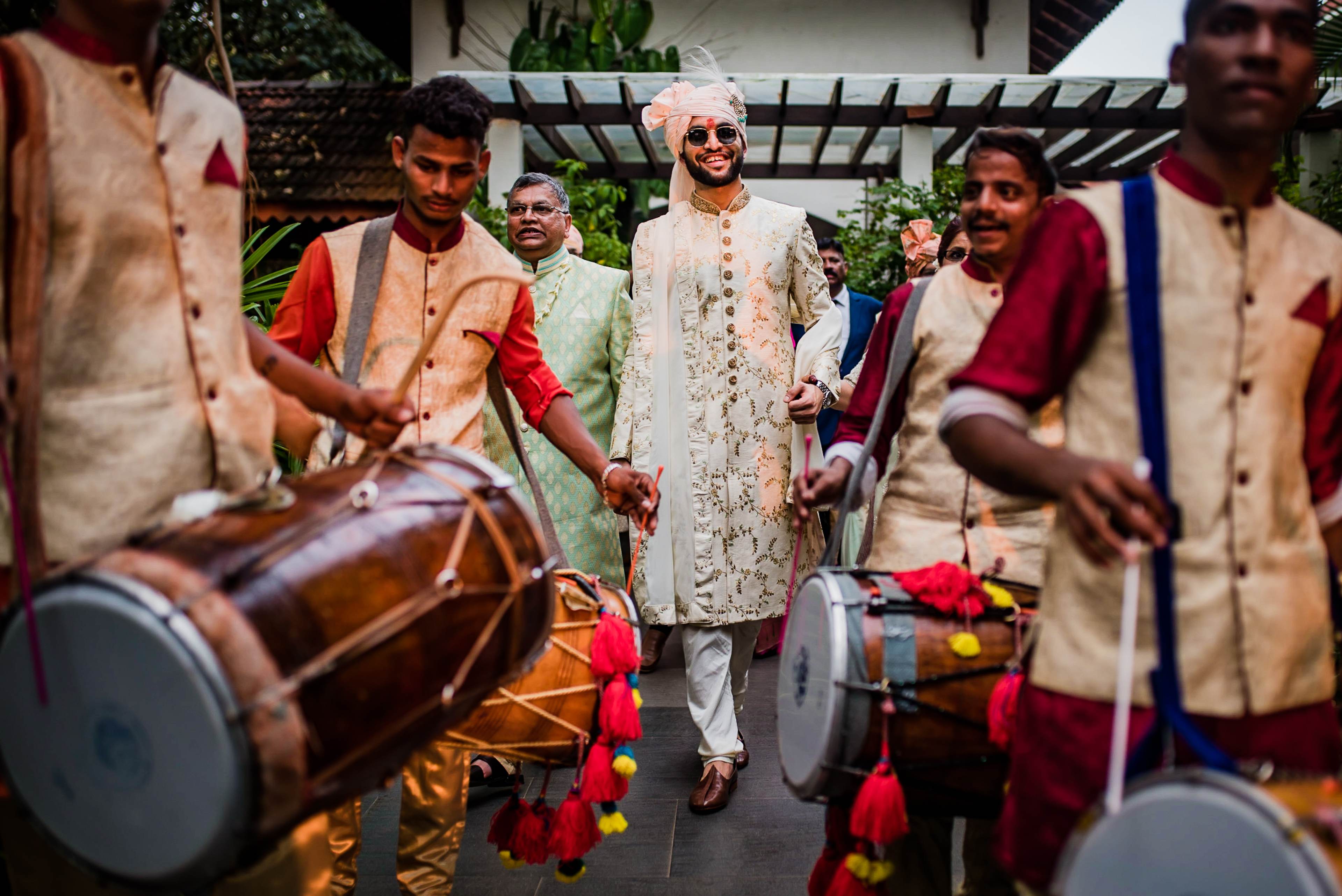 Avani and Srikant walk through a festive garden ceremony in Goa, surrounded by drummers in traditional attire