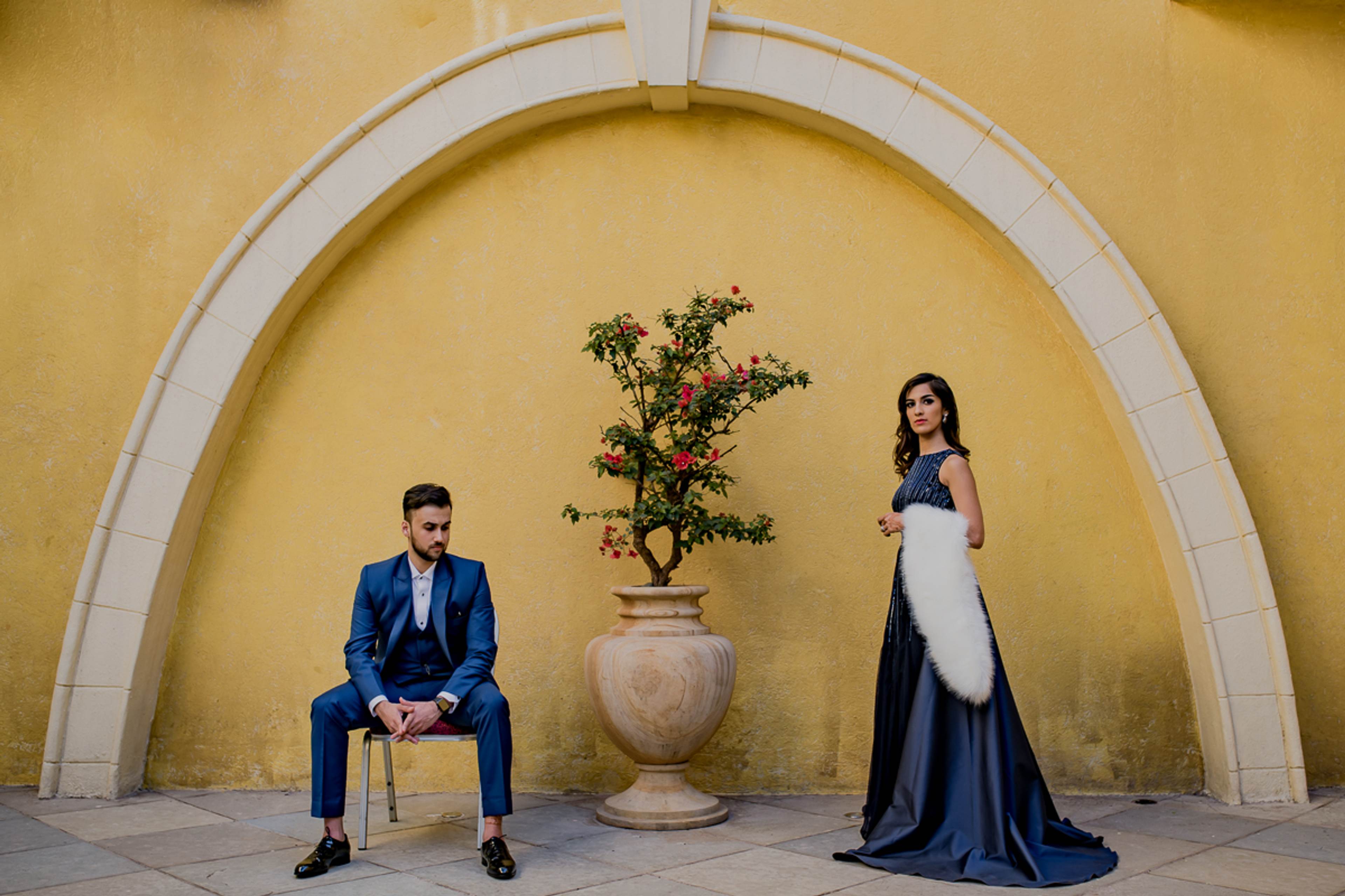 Man in blue suit seated on chair, woman in white and navy gown standing, potted plant between them against yellow arched wall
