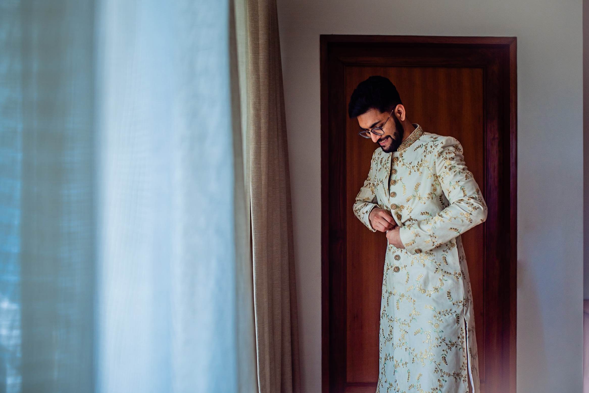 Groom Srikant in cream embroidered sherwani, adjusting cufflinks in doorway before wedding