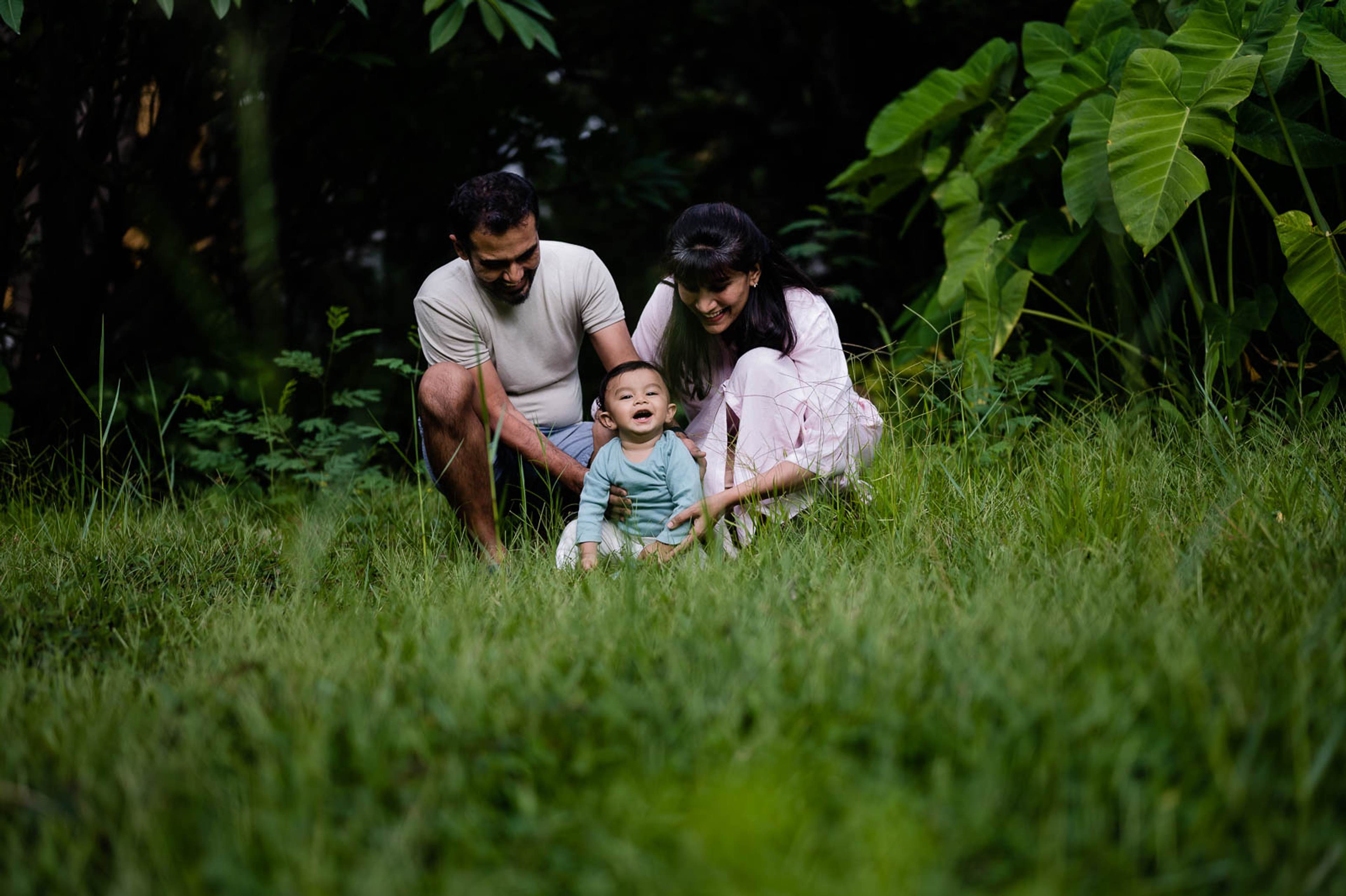 Two adults and a toddler in blue shirt playing in lush green grass surrounded by foliage