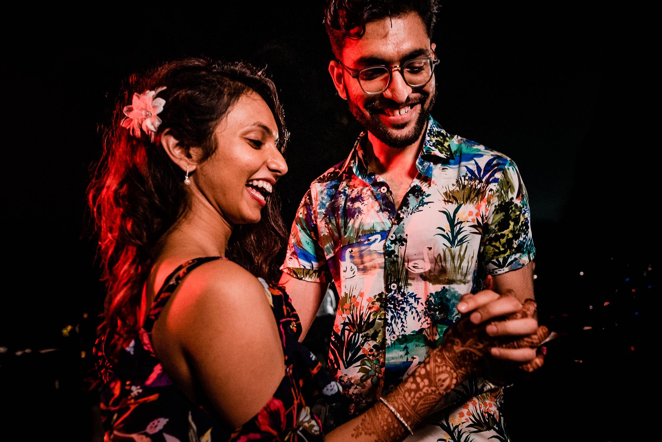 Avani and Srikant laugh together in warm red light, she wearing a flower in her hair, he in a colorful floral shirt, Goa