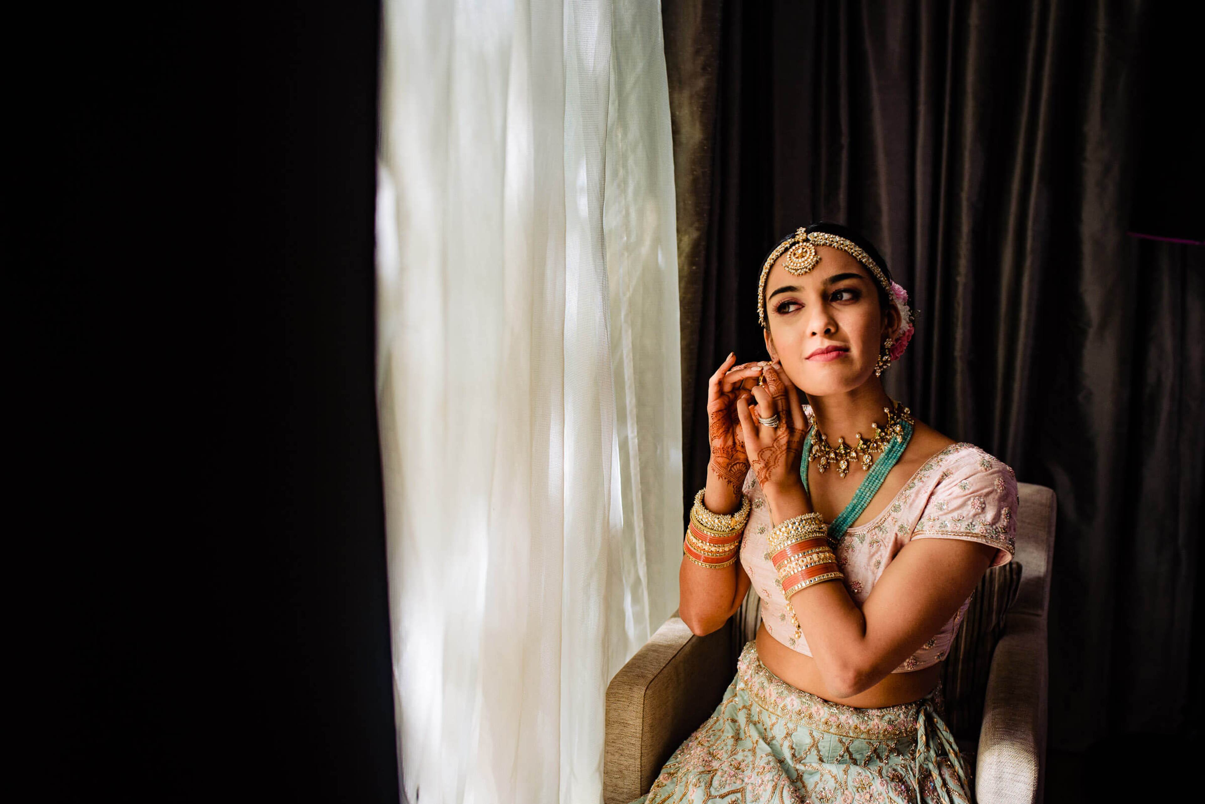 Shreya seated by window light in traditional bridal attire, jewelry and henna, thoughtful expression