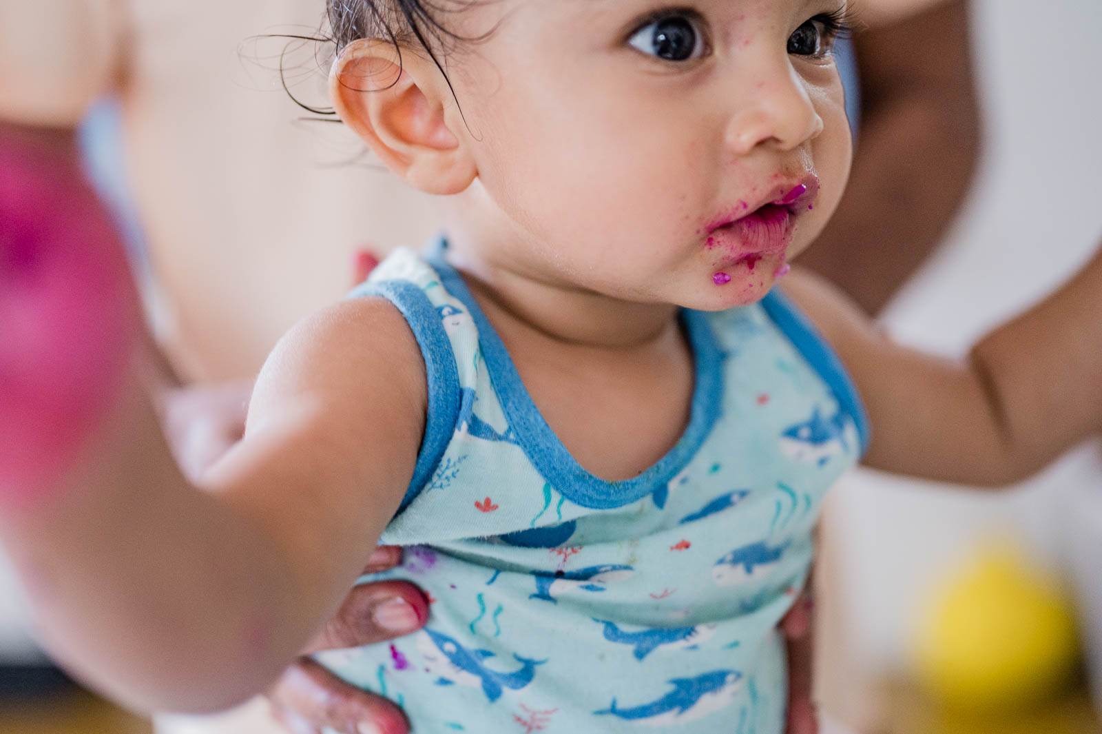Toddler in blue patterned tank top with pink-stained mouth, held by caregiver indoors
