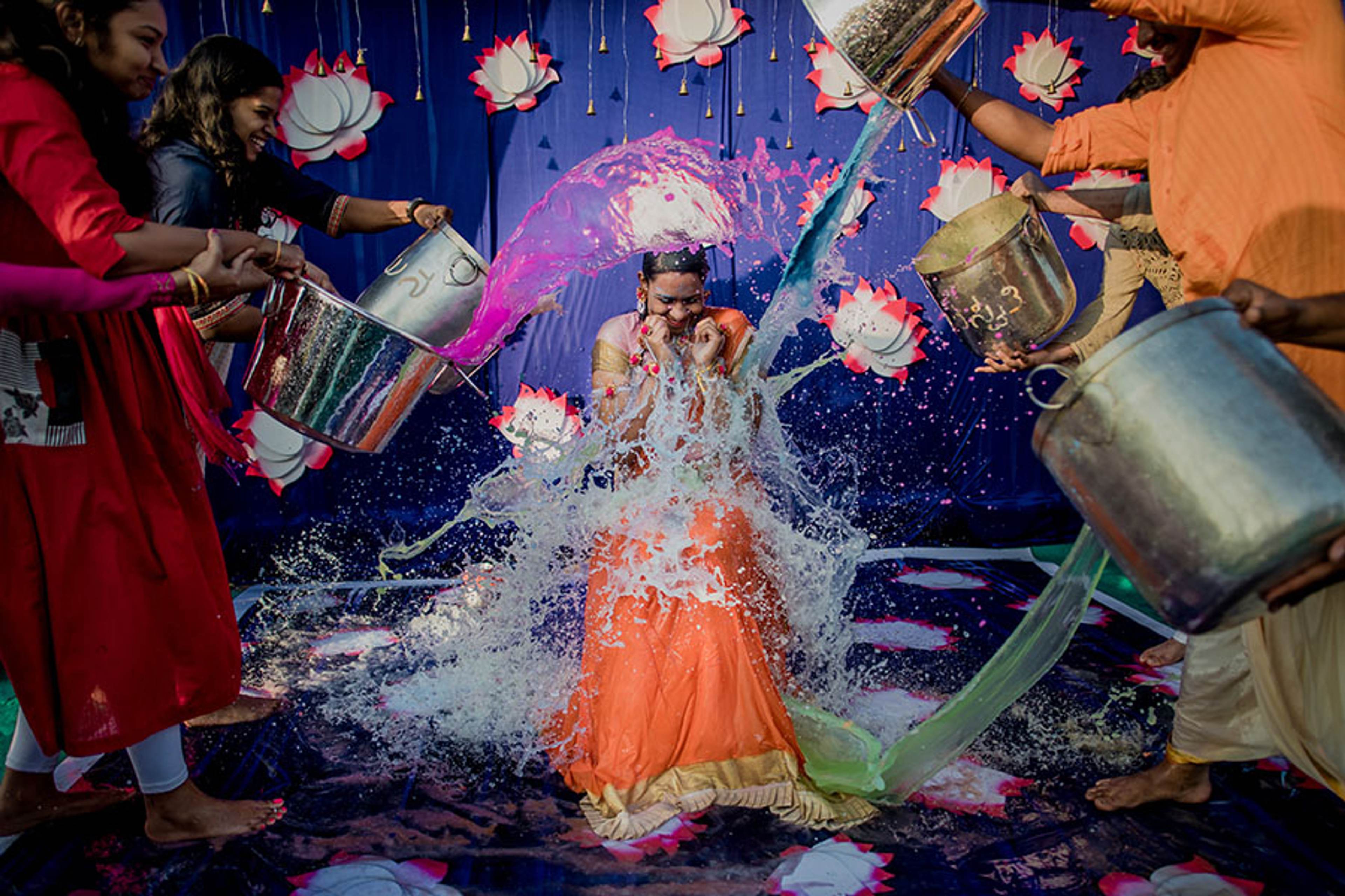 Woman in orange dress splashed with water by guests pouring buckets against blue floral backdrop