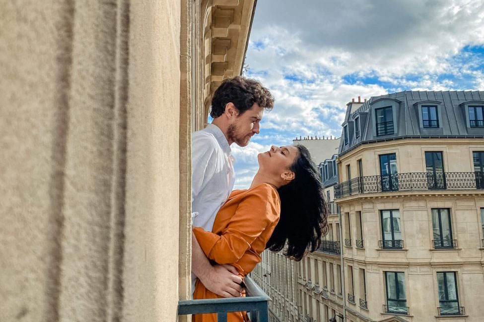 Couple embracing on a Parisian balcony with classic architecture and blue sky behind them by Rimi Sen