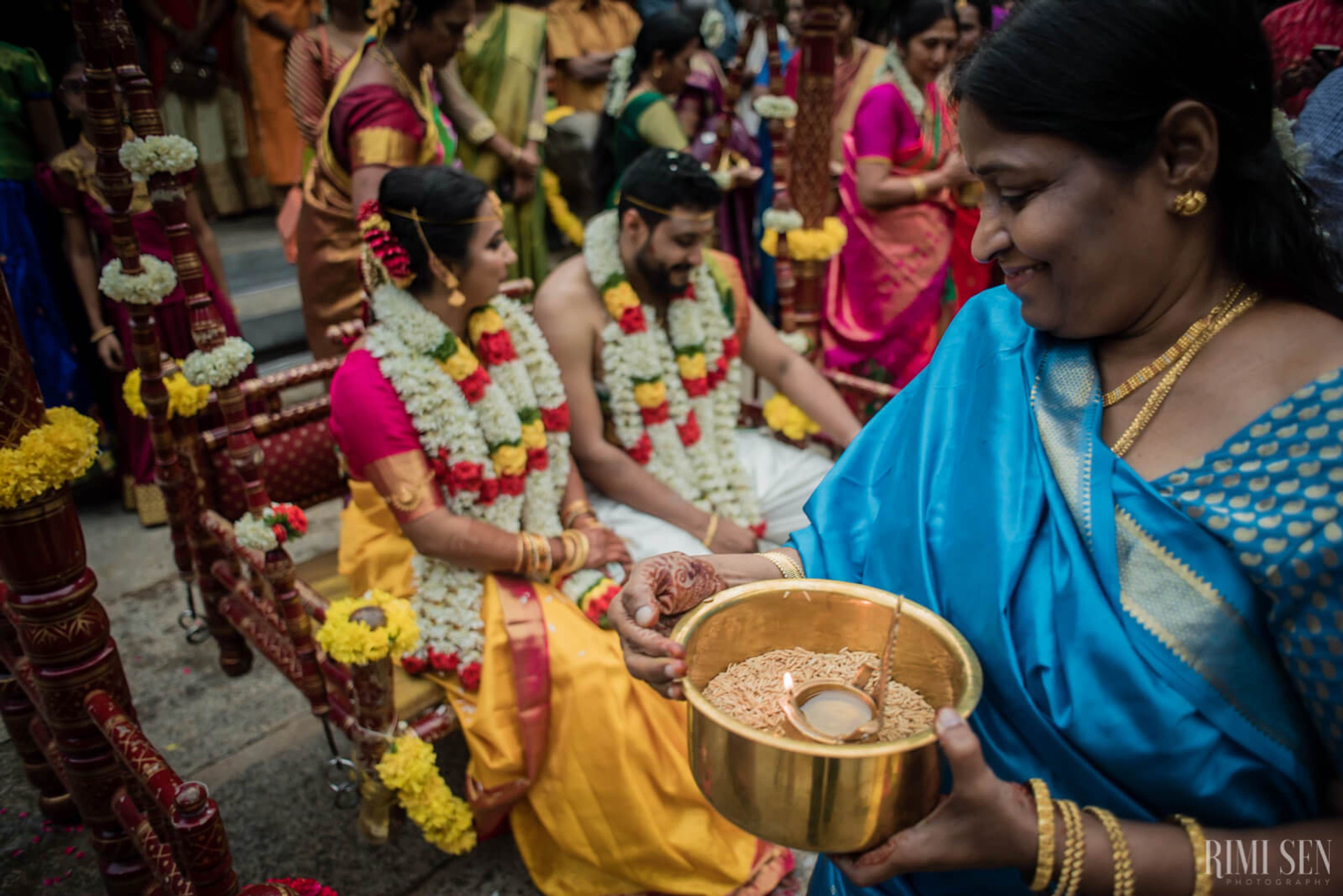 Woman in blue saree offers ceremonial rice to couple adorned with flower garlands during Bangalore wedding