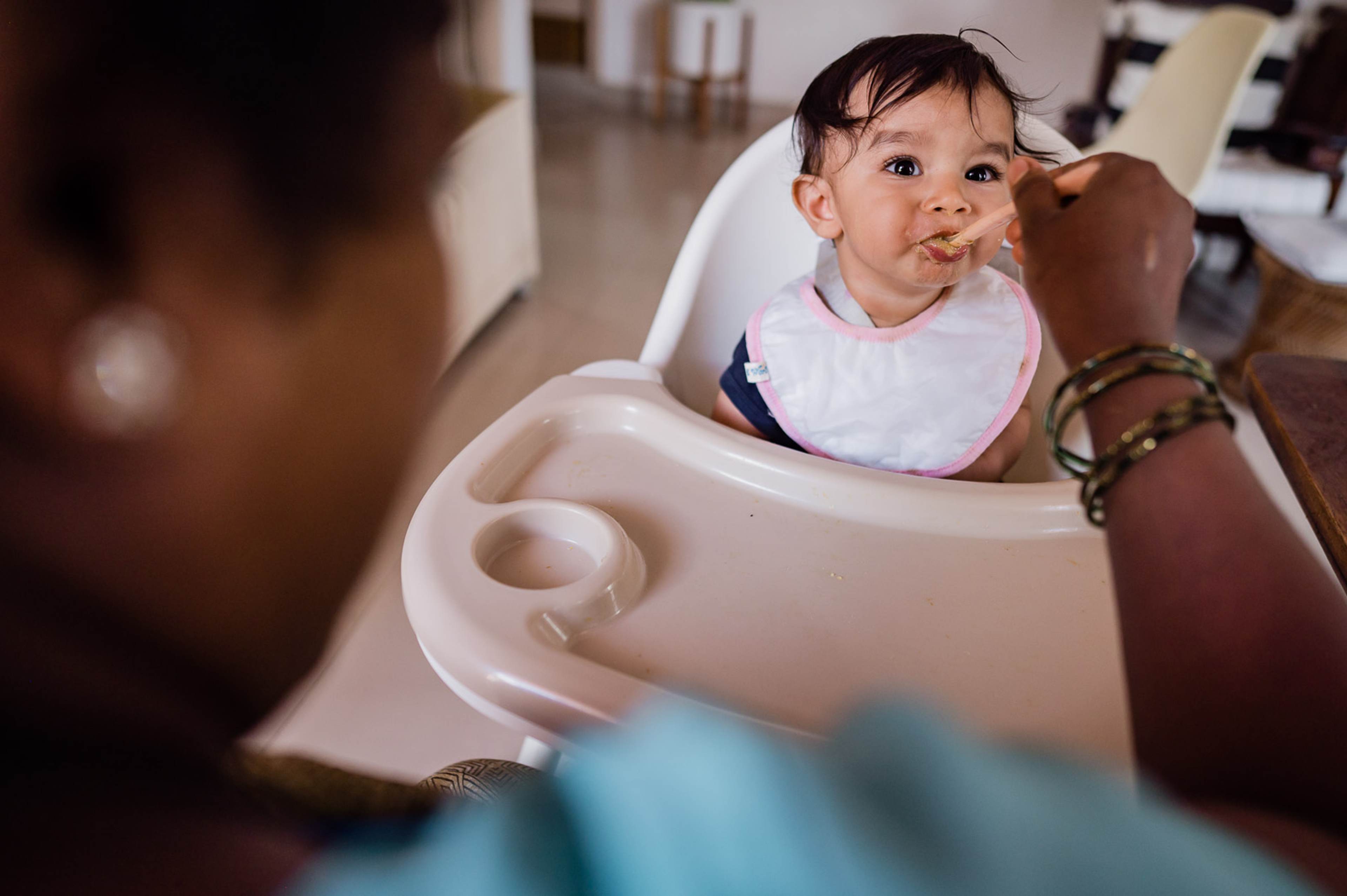 Toddler in white bib at high chair, adult feeding in warm indoor light