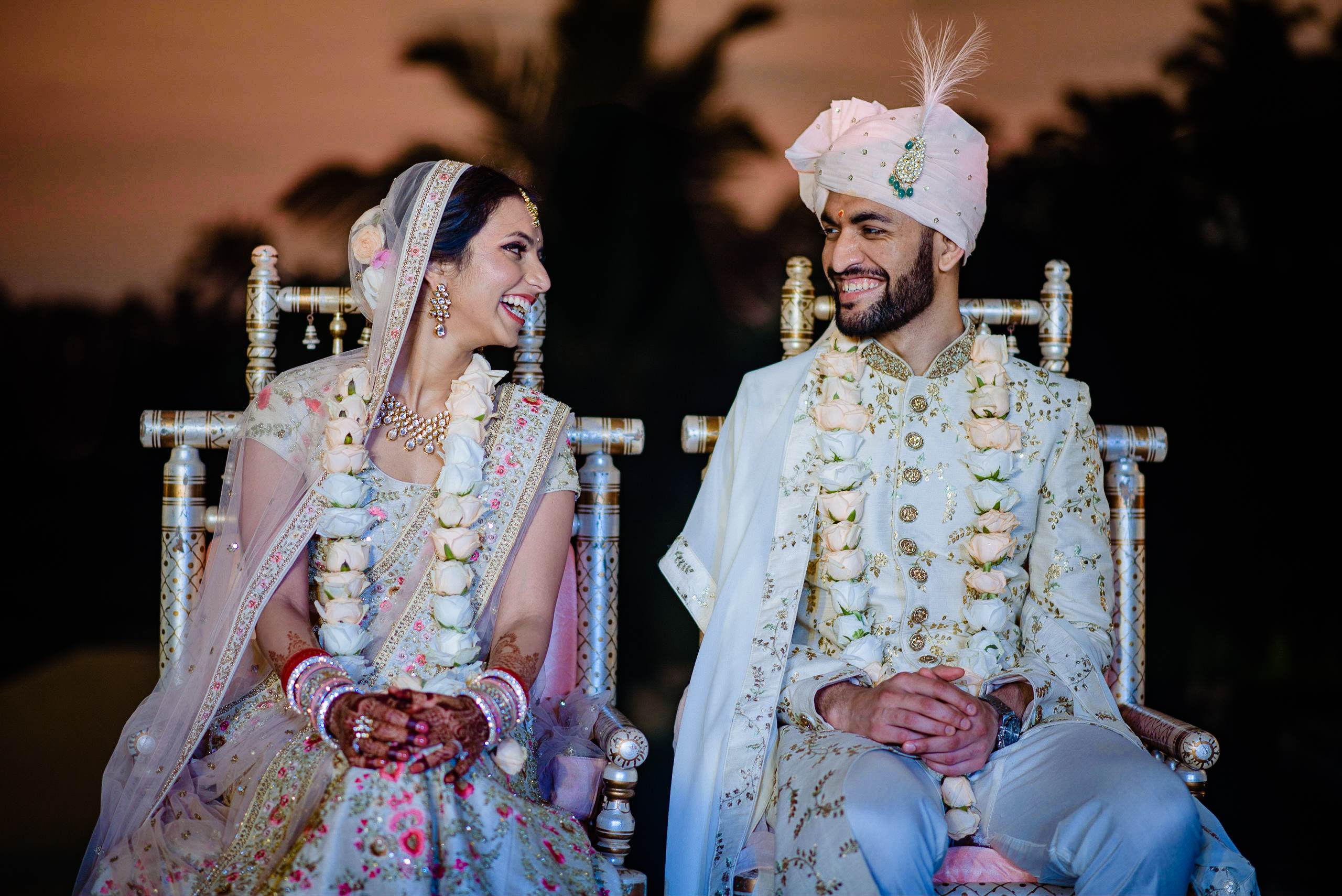 Avani and Srikant sit together in ornate chairs at their Goa wedding, dressed in traditional Indian attire, smiling at each other