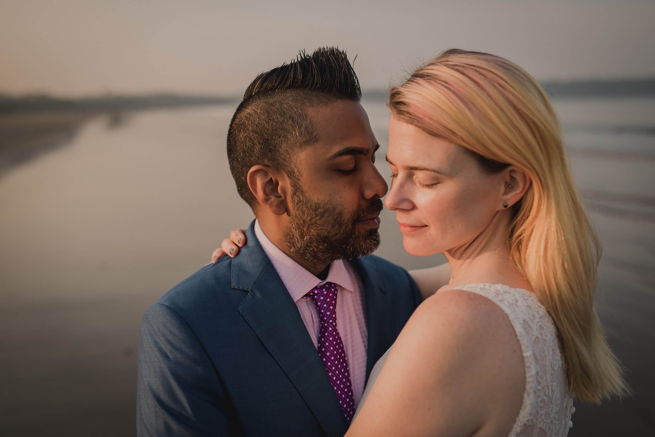 Sarah and Ravin embrace at sunset by calm waters in Goa, soft golden light on their faces
