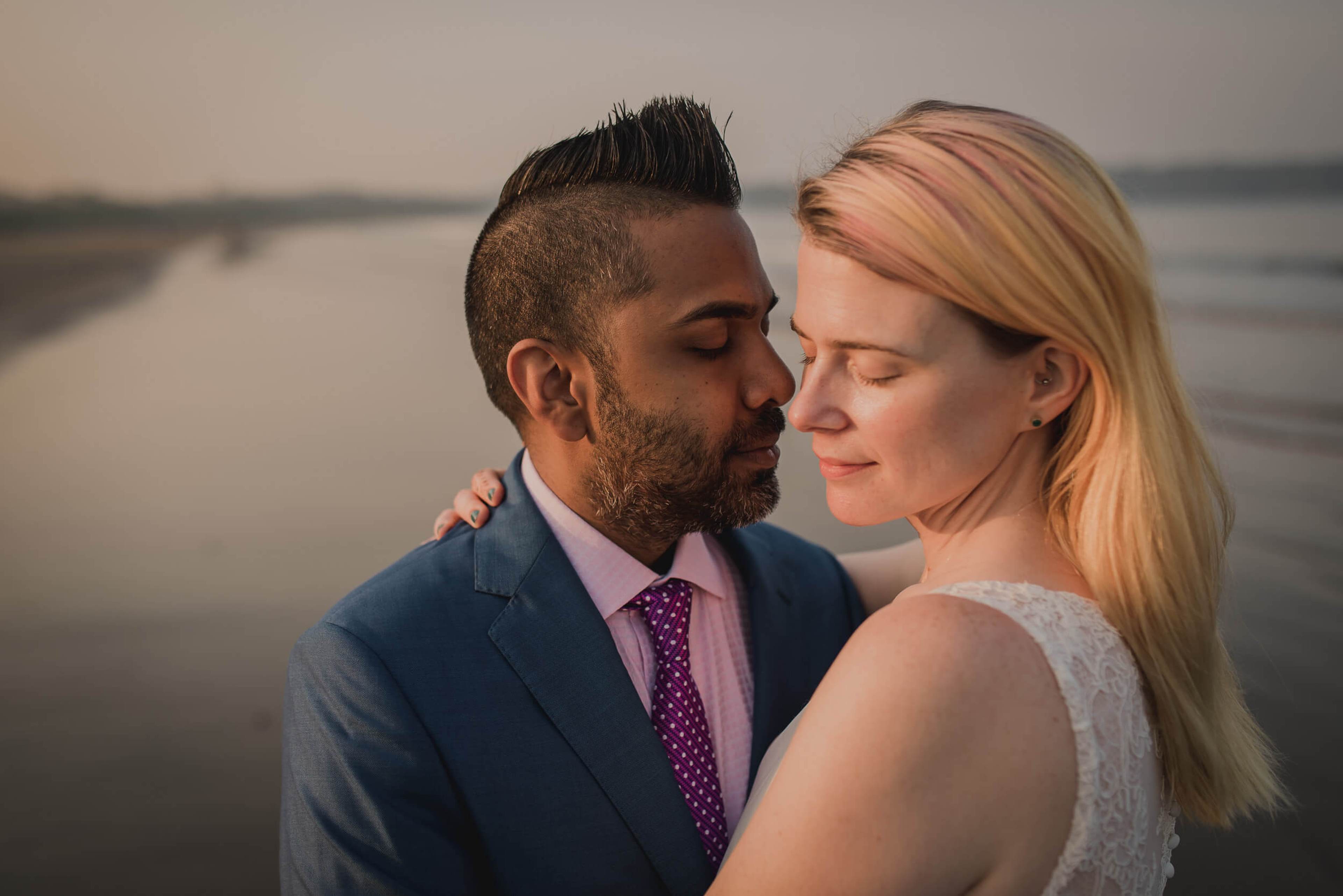 Sarah and Ravin embrace at sunset by calm waters in Goa, soft golden light on their faces