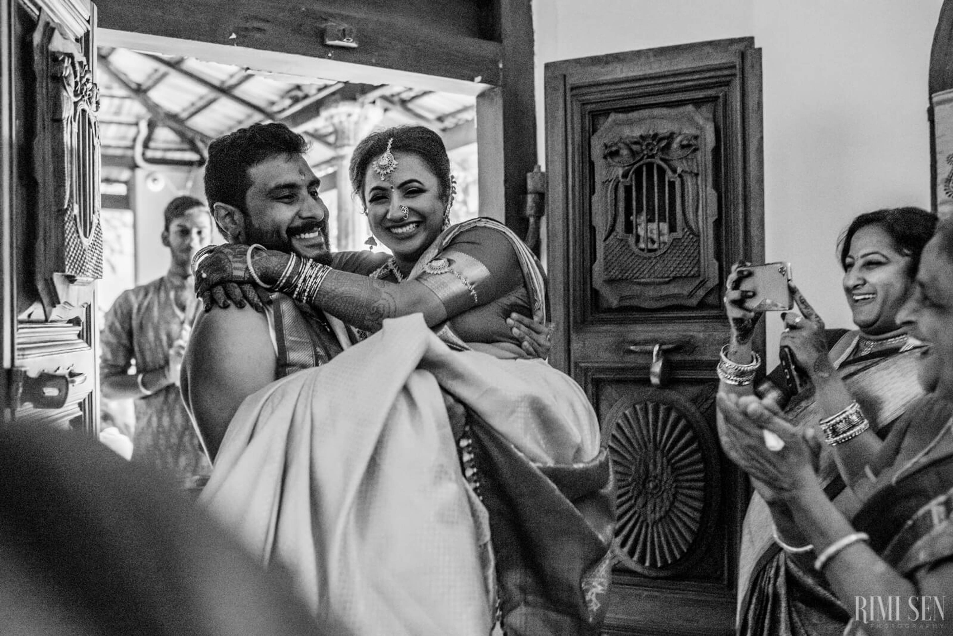 Groom carries bride through ornate doorway as guests celebrate and photograph the moment in black and white