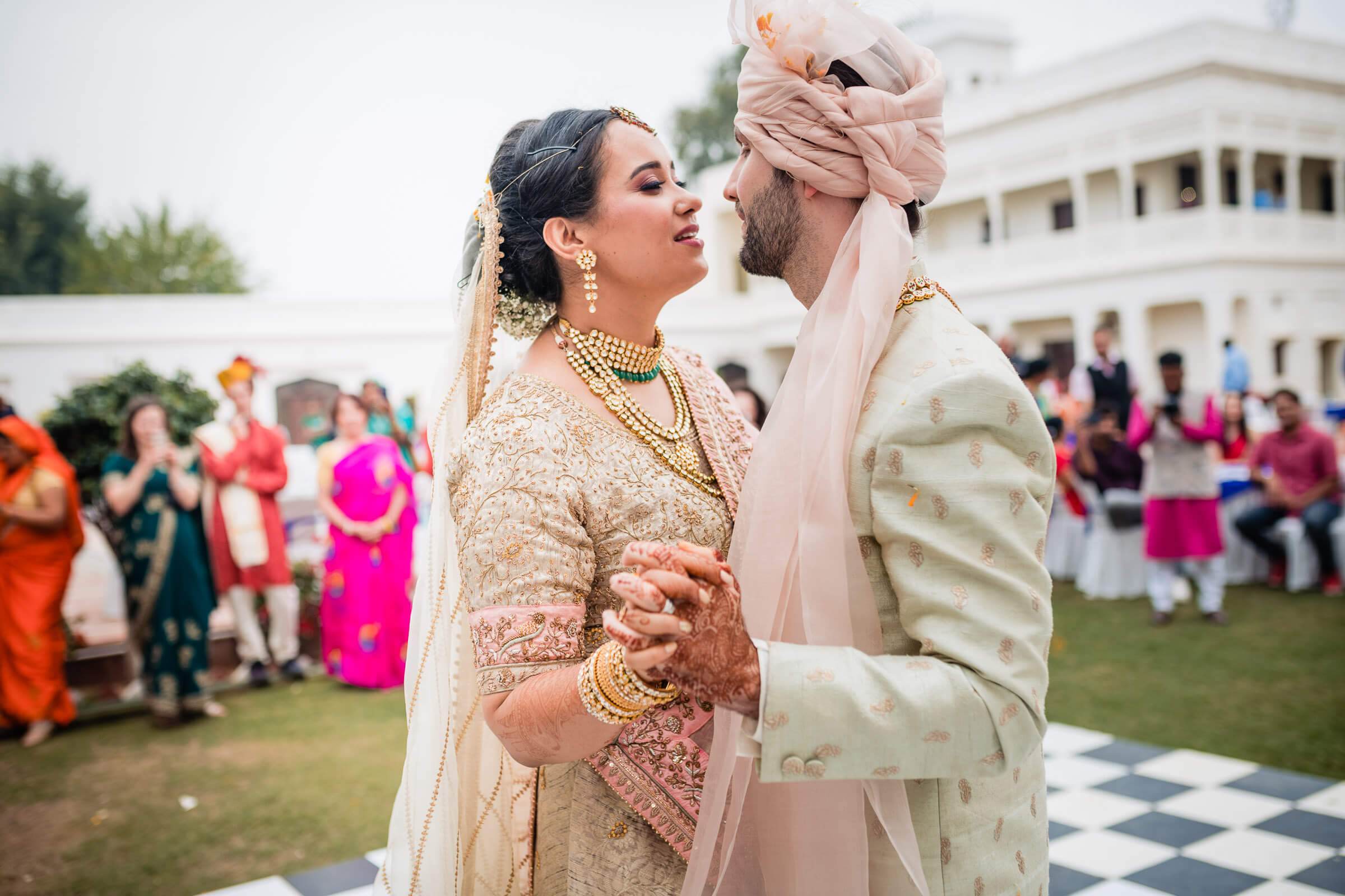 Maitrayee and Eric embrace in ornate traditional attire at their Rajasthan wedding celebration, guests dancing in vibrant saris behind them