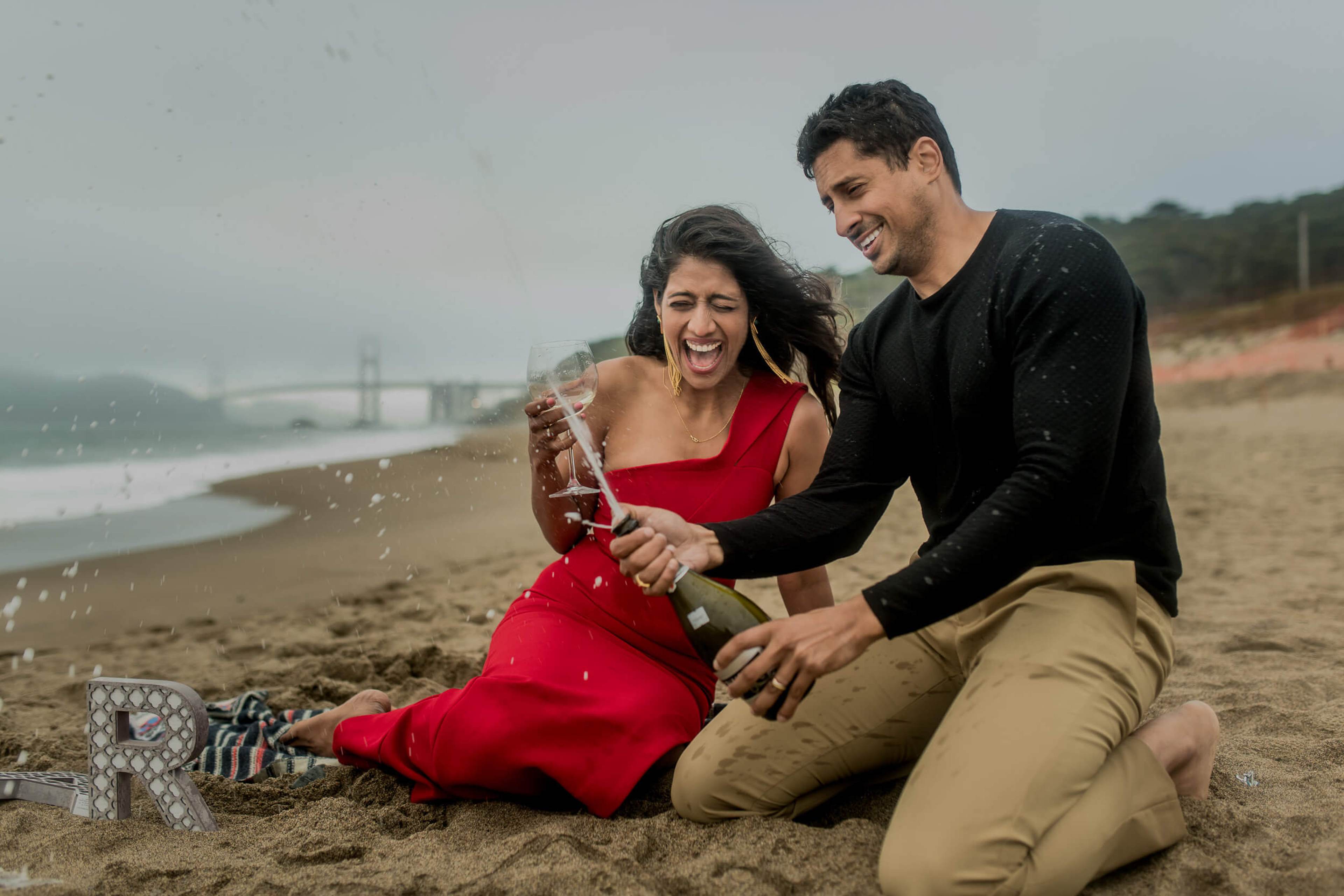 Anu and Raghu pop champagne on a California beach at sunset, Golden Gate Bridge visible in the distance