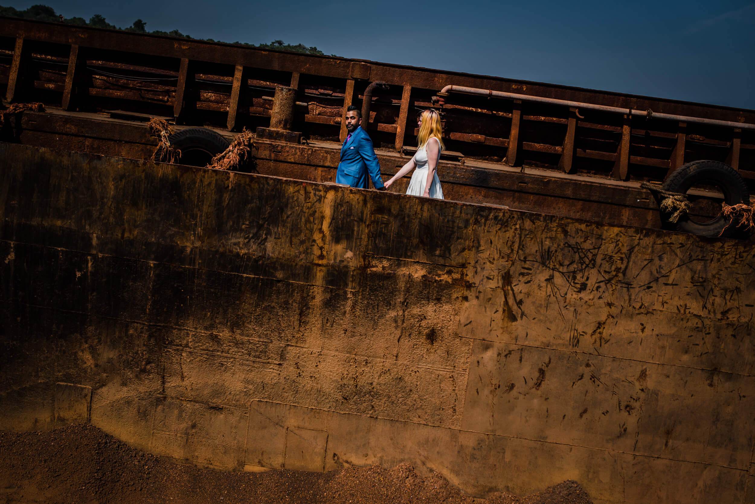 Sarah and Ravin walk holding hands along a weathered shipwreck in Goa at dusk