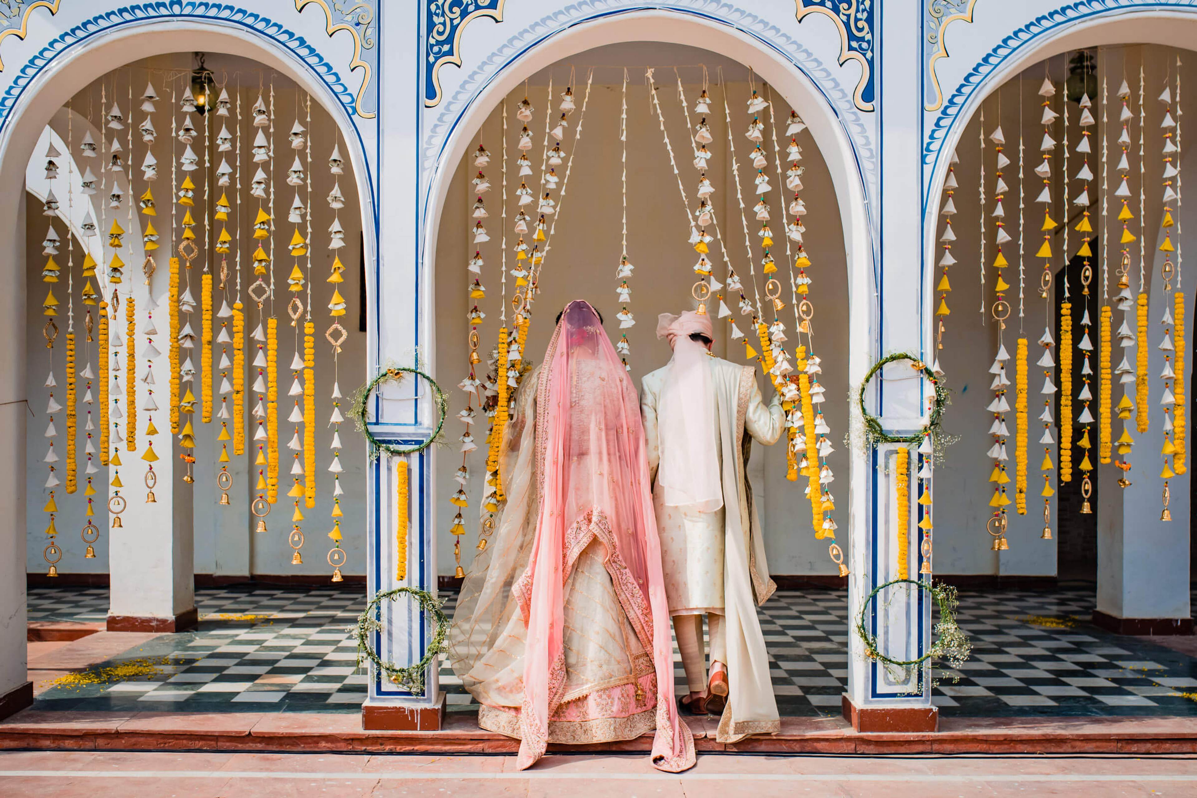 Maitrayee in pink veil and Eric in cream sherwani beneath hanging marigold garlands, Rajasthan