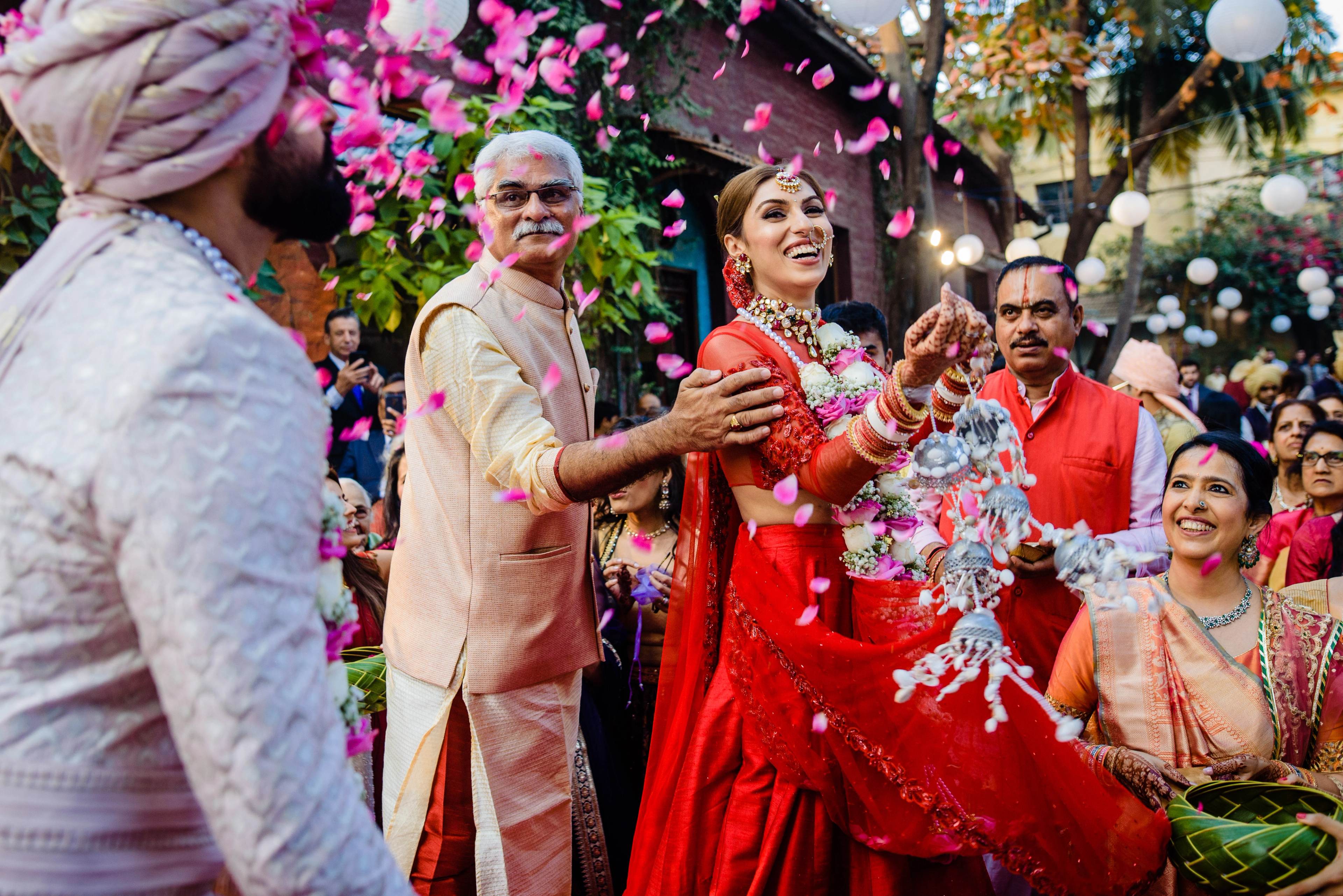 Woman in red embellished lehenga celebrates with family amid vibrant pink floral backdrop and petals