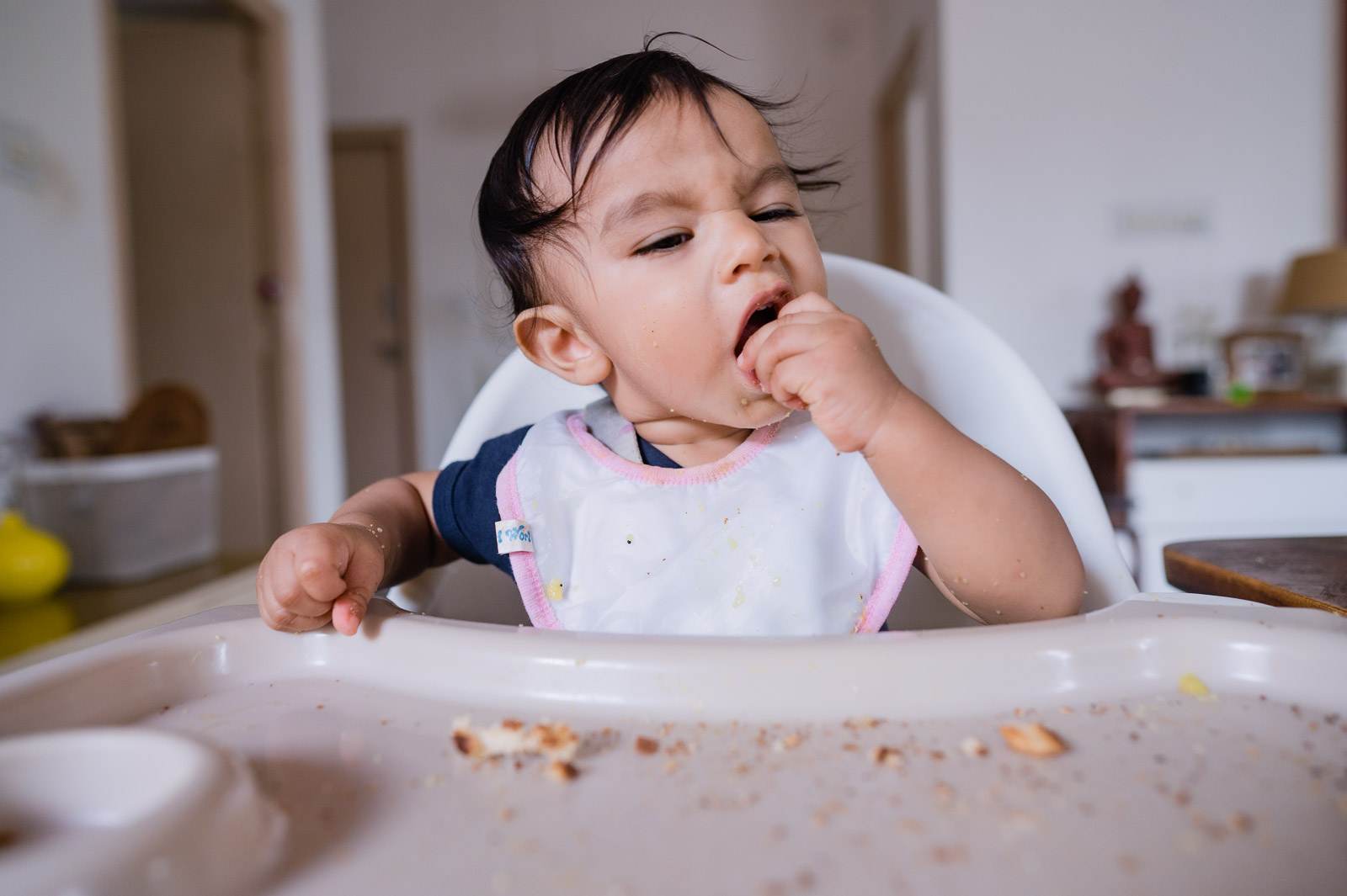 Toddler in high chair eating messily, hand to mouth, scattered crumbs on tray