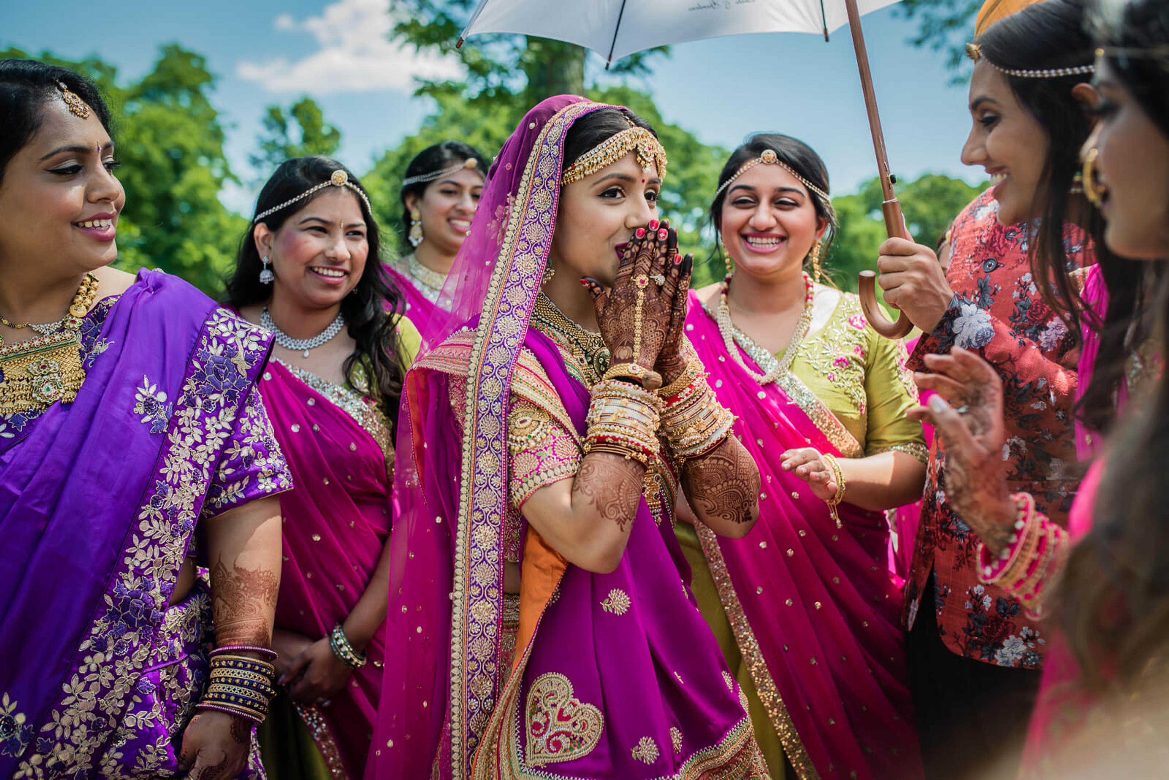 Barkha surrounded by bridesmaids in vibrant traditional attire and henna, laughing under umbrella outdoors