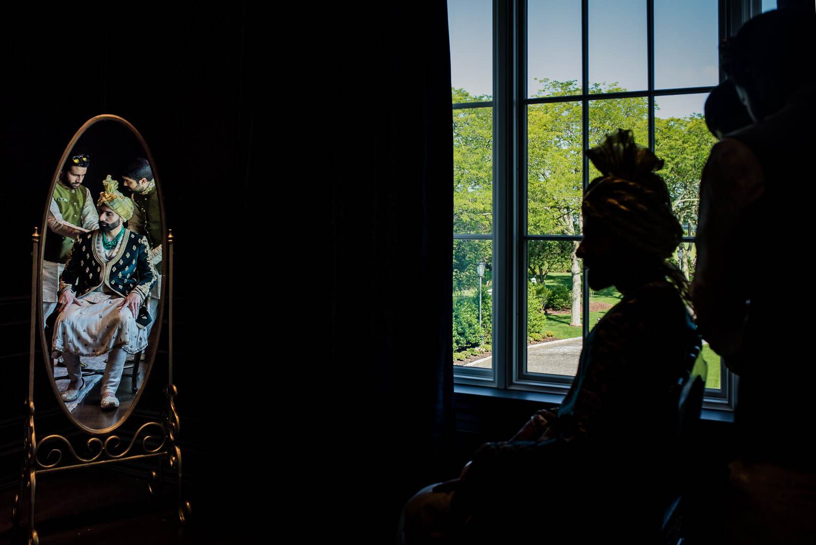 Barkha and Ankit reflected in ornate mirror, silhouetted figures by sunlit window overlooking green landscape