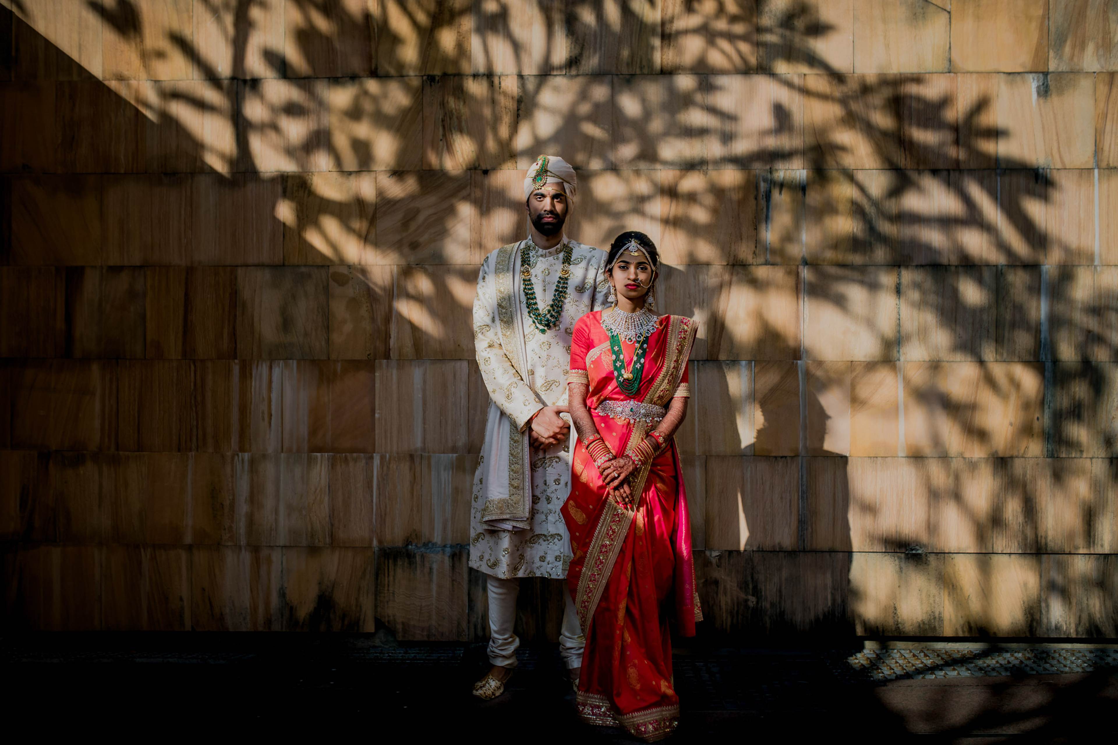 Couple in traditional Indian wedding attire standing against wooden wall with dappled tree shadows