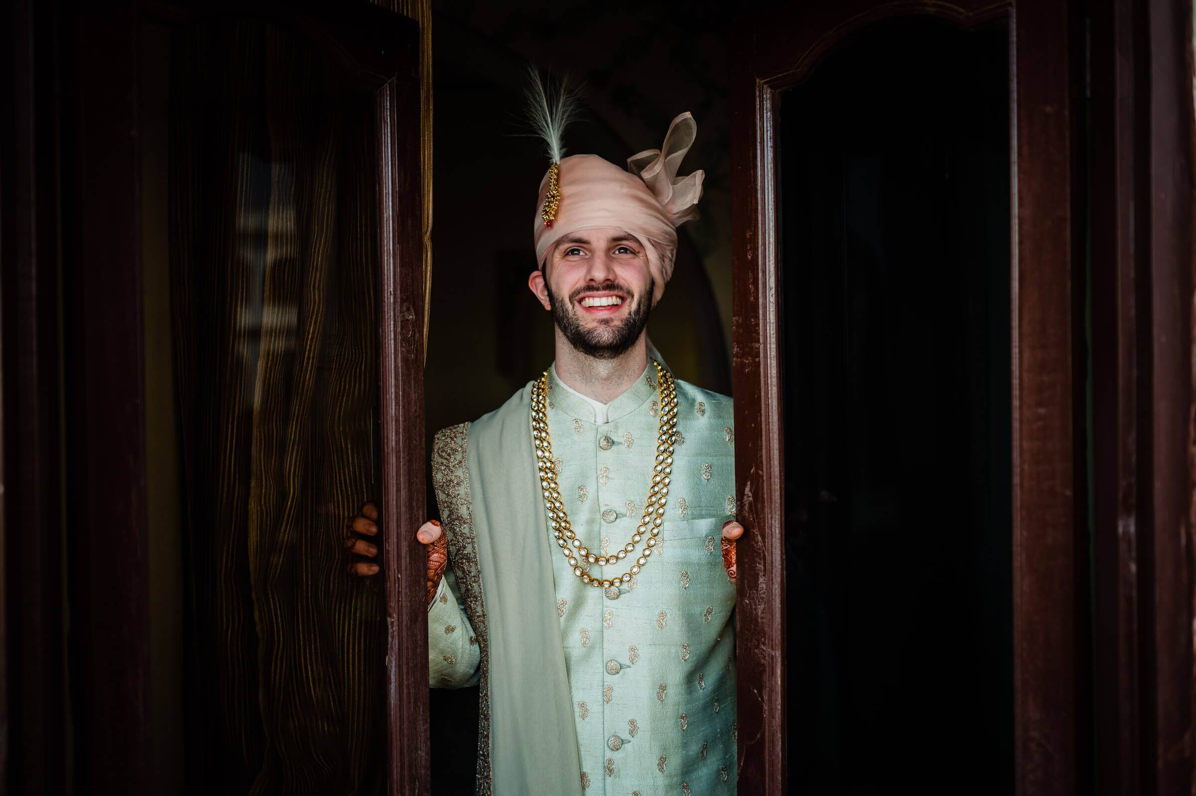 Eric stands smiling in doorway wearing pale green sherwani with gold chain and feathered turban, Rajasthan