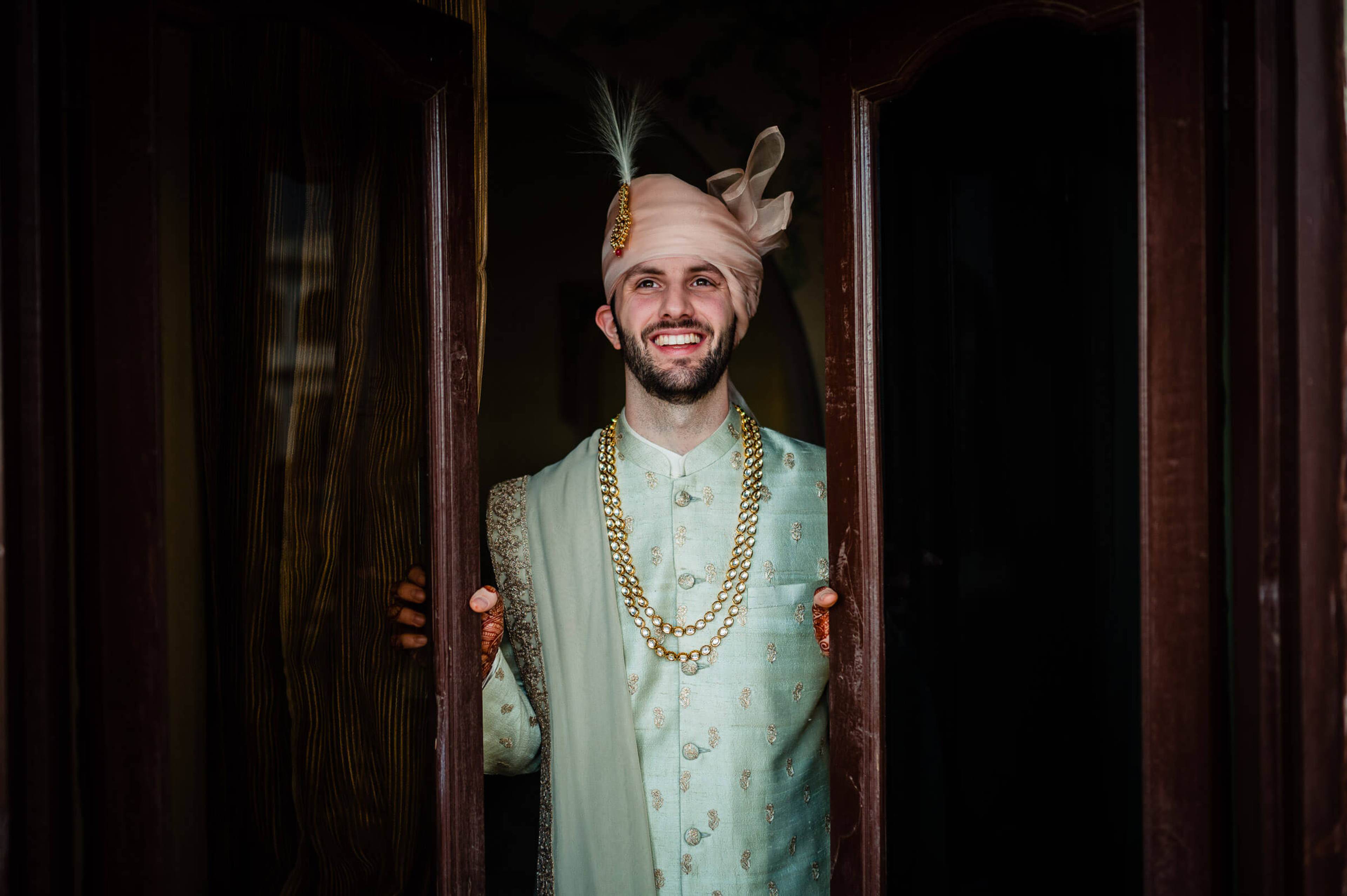 Eric stands smiling in doorway wearing pale green sherwani with gold chain and feathered turban, Rajasthan