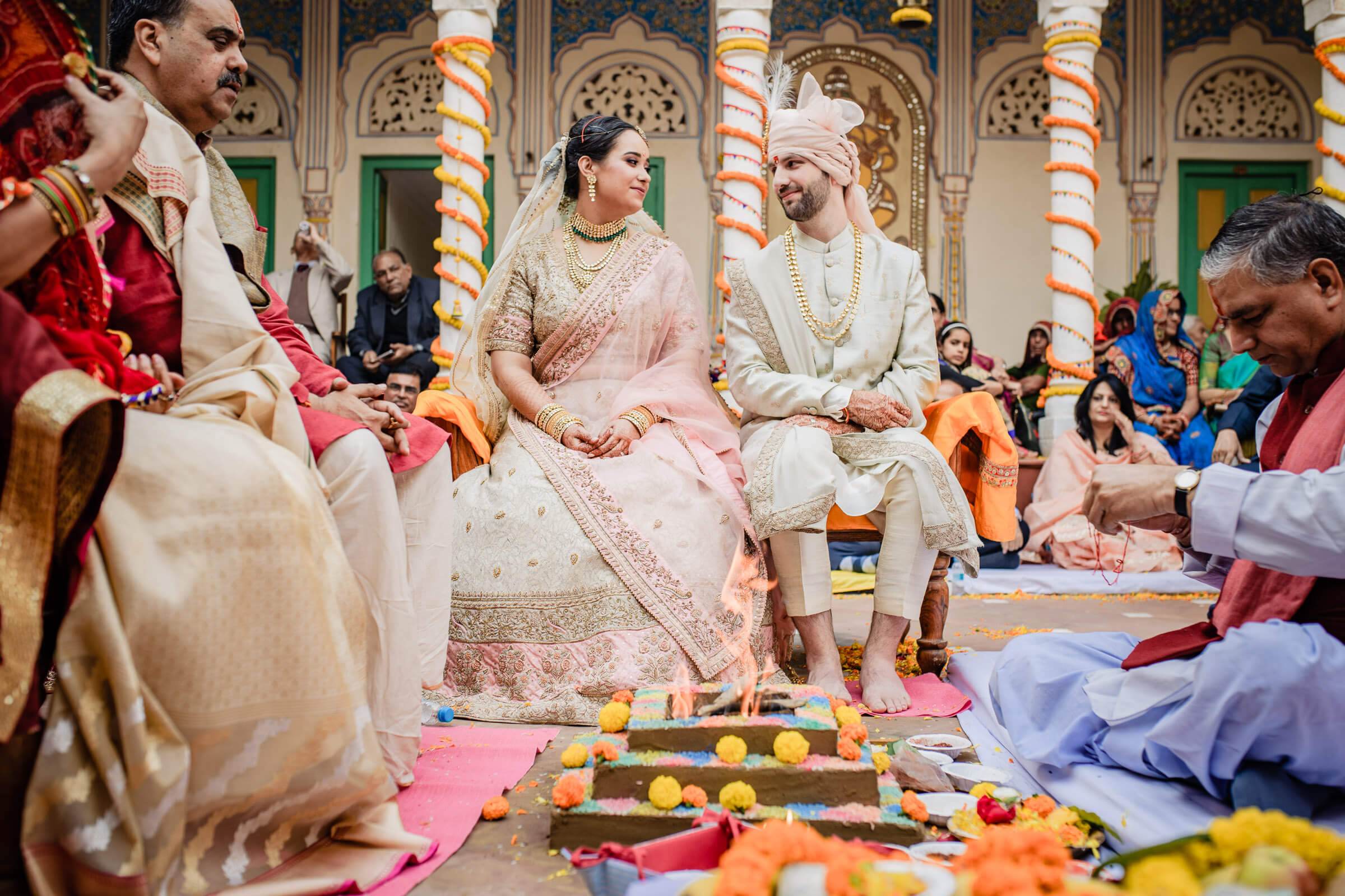 Maitrayee and Eric sit together during an ornate temple ceremony in Rajasthan, surrounded by family and floral decorations