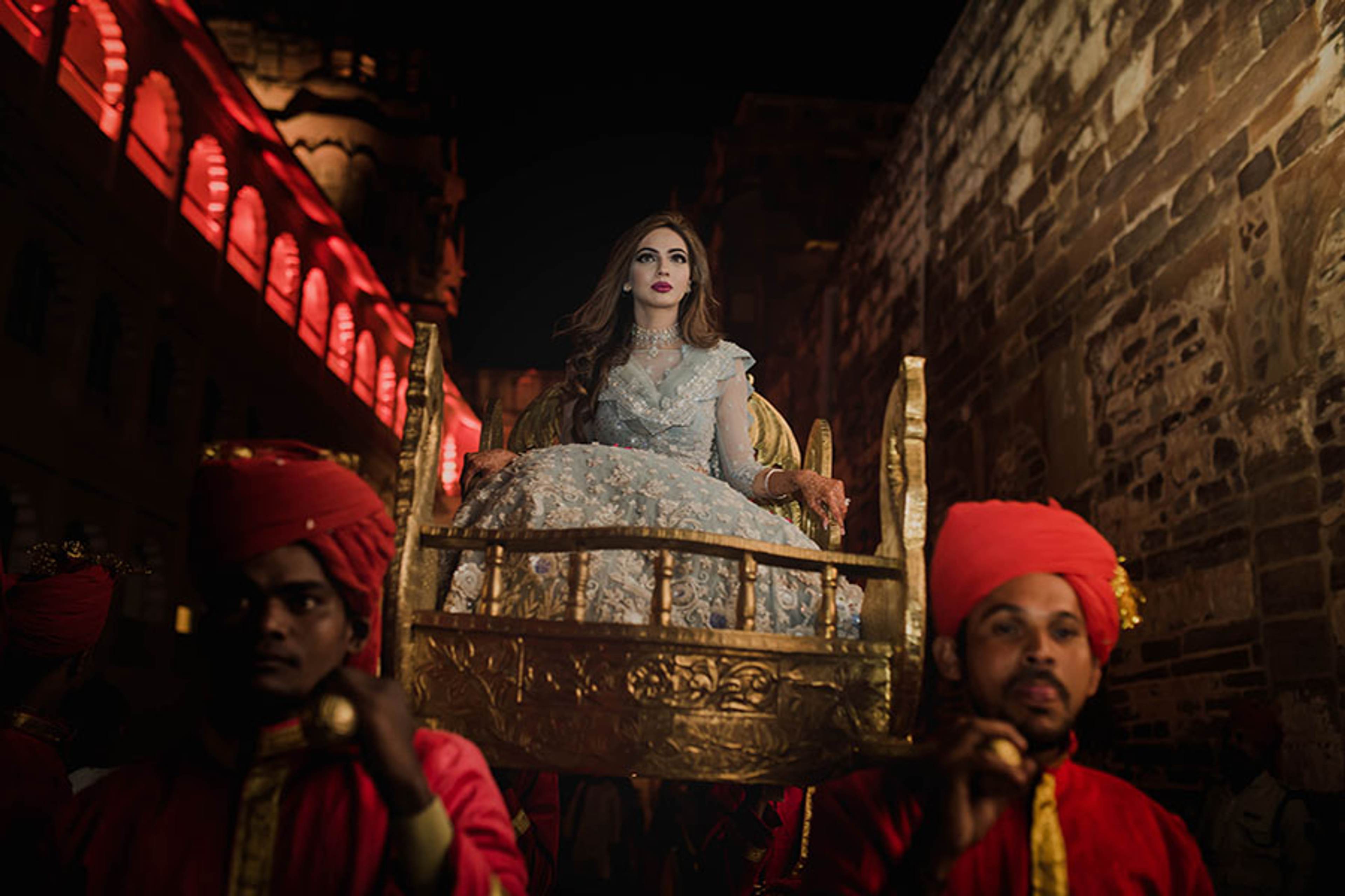 Woman in embellished gown on ornate golden bed, men in red turbans beside her in candlelit stone setting
