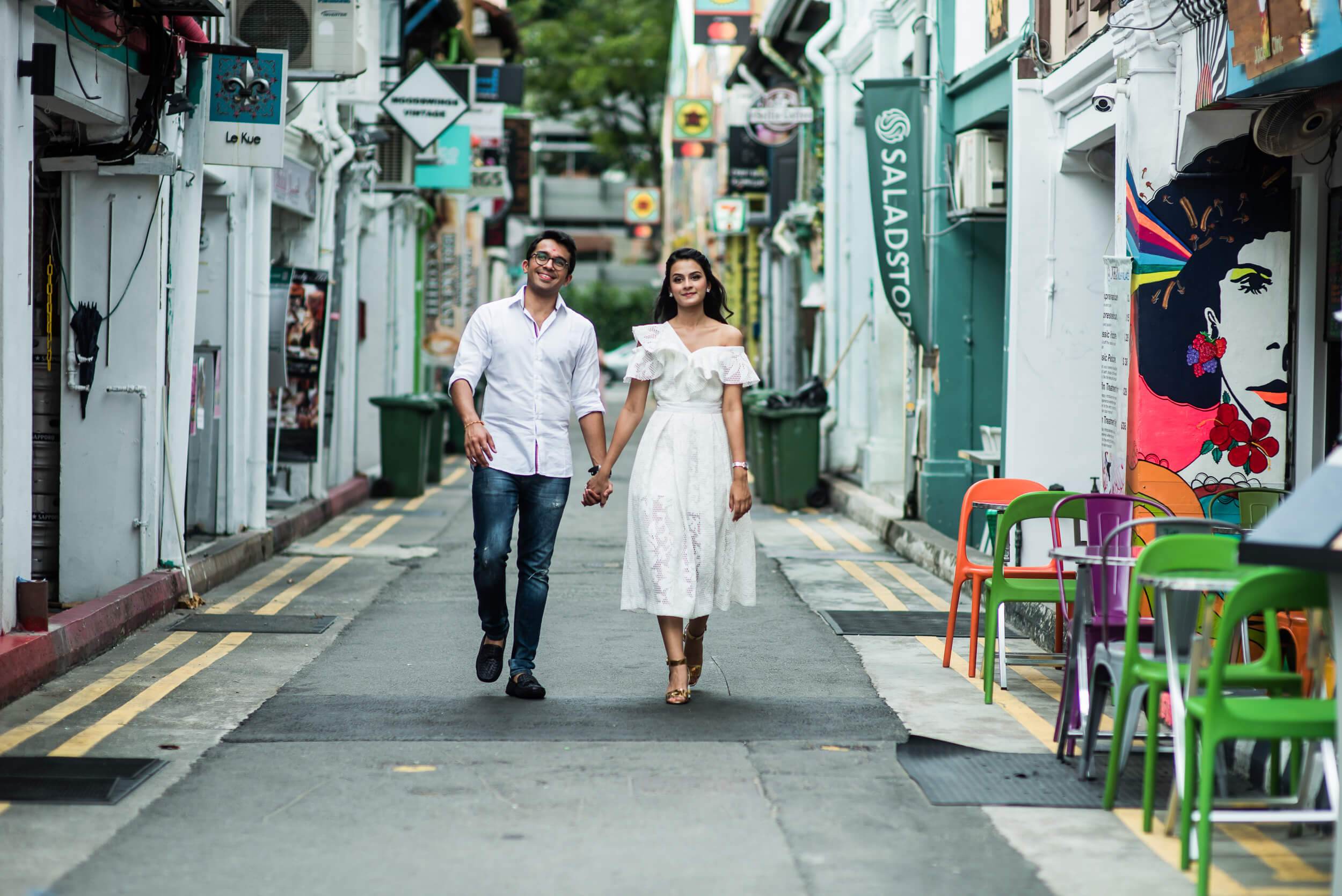 Yash and Disha hold hands walking down a vibrant Singapore alley lined with colorful shops