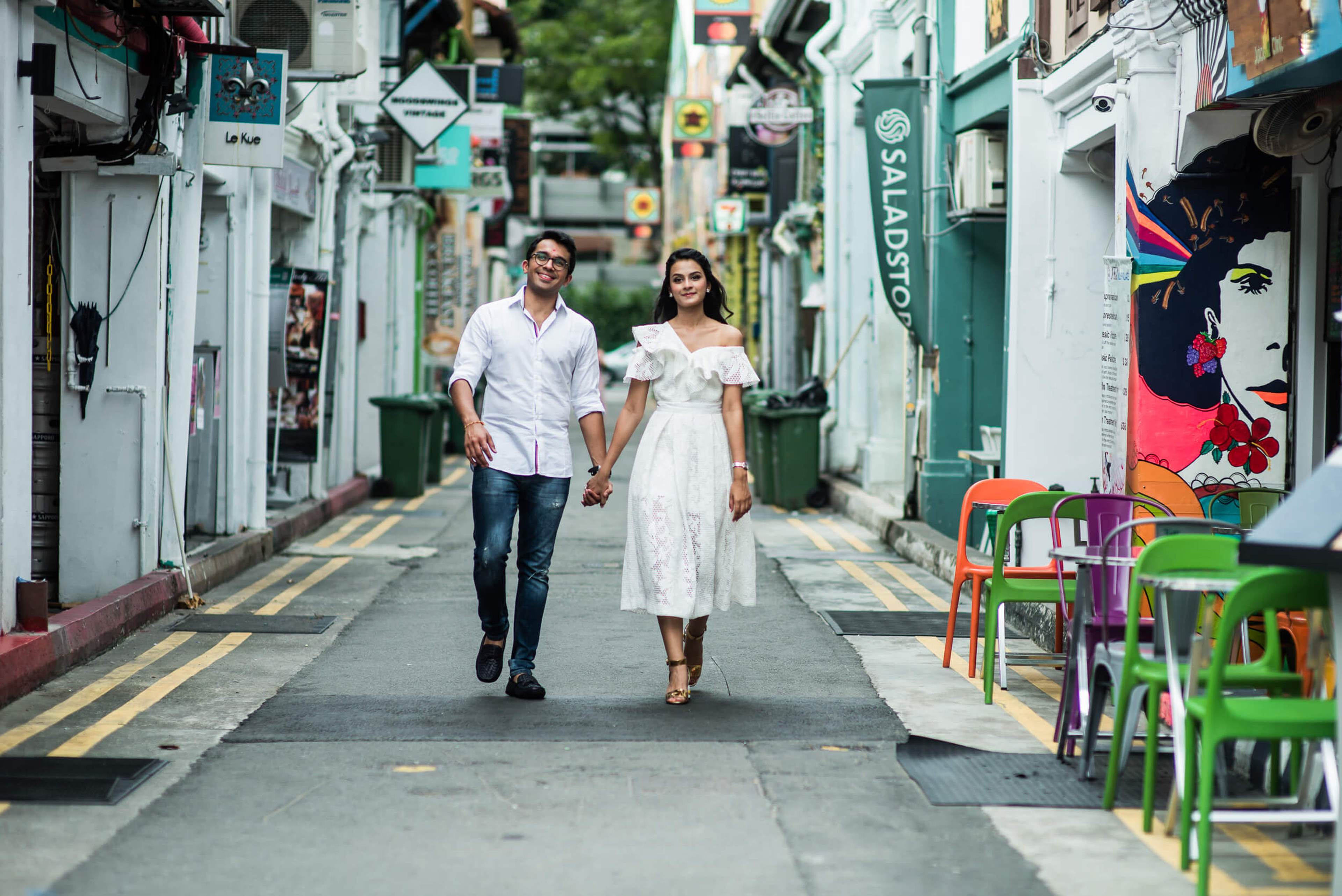Yash and Disha hold hands walking down a vibrant Singapore alley lined with colorful shops